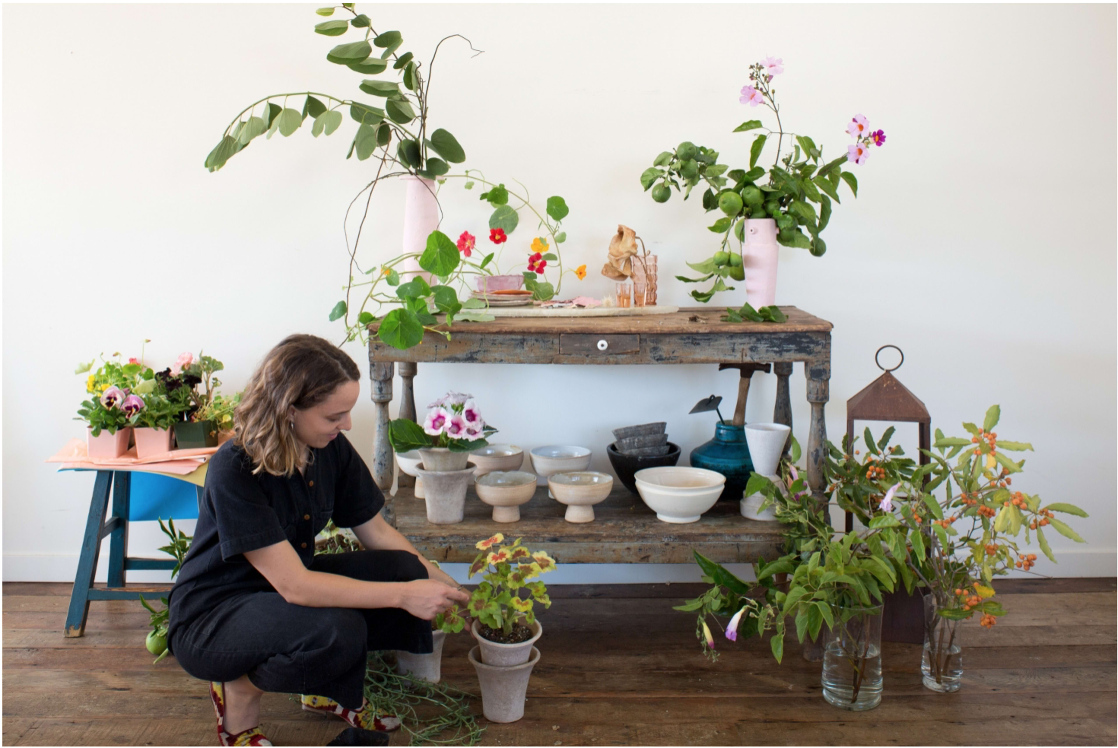 A woman tends to plants on a wooden shelf adorned with various pots and flowers against a plain wall.