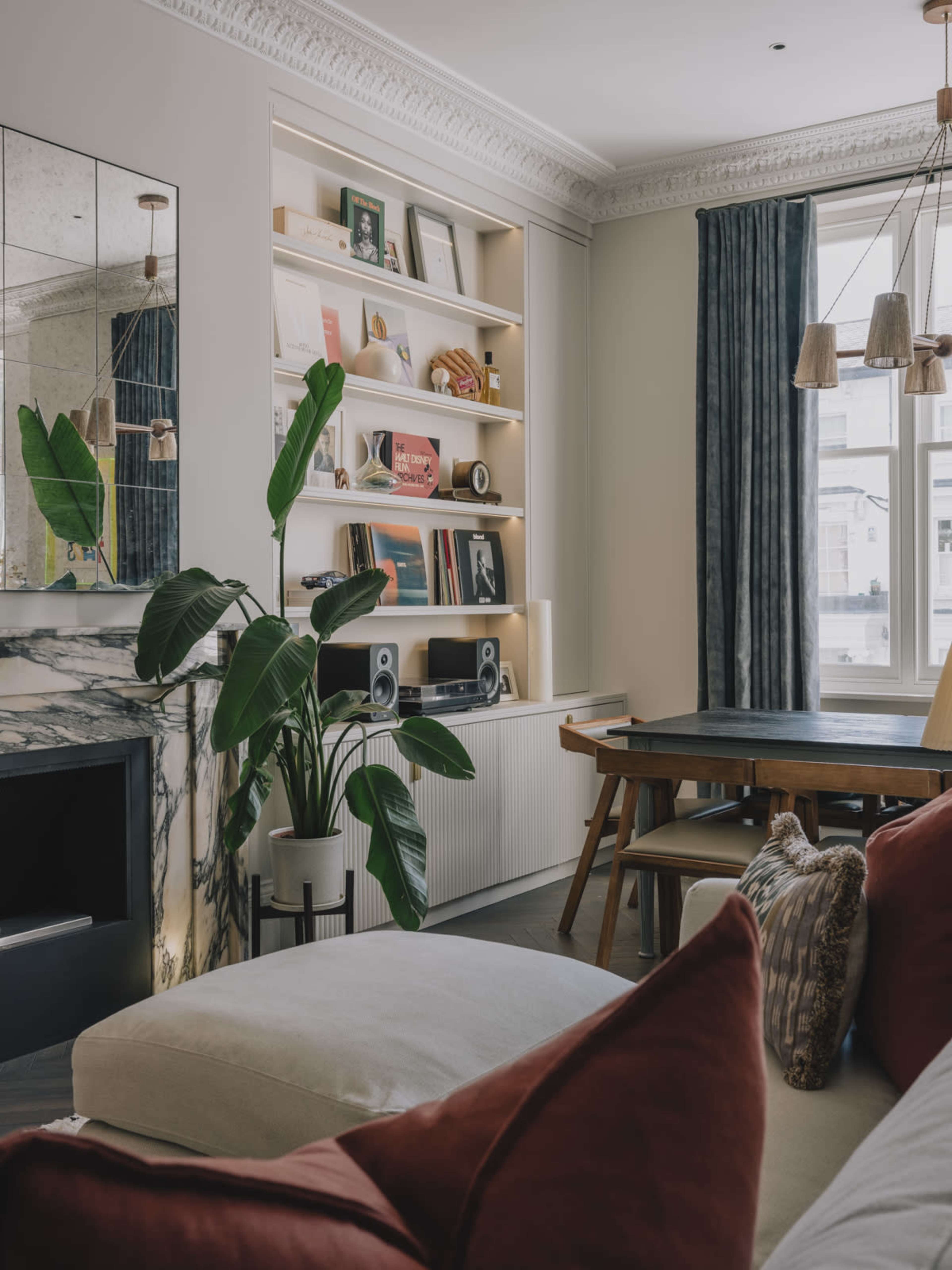 A modern living room features a white sofa with red cushions, a wooden dining table, a fireplace with marble surrounding, shelves filled with books and decor, potted plants, and large windows with curtains.