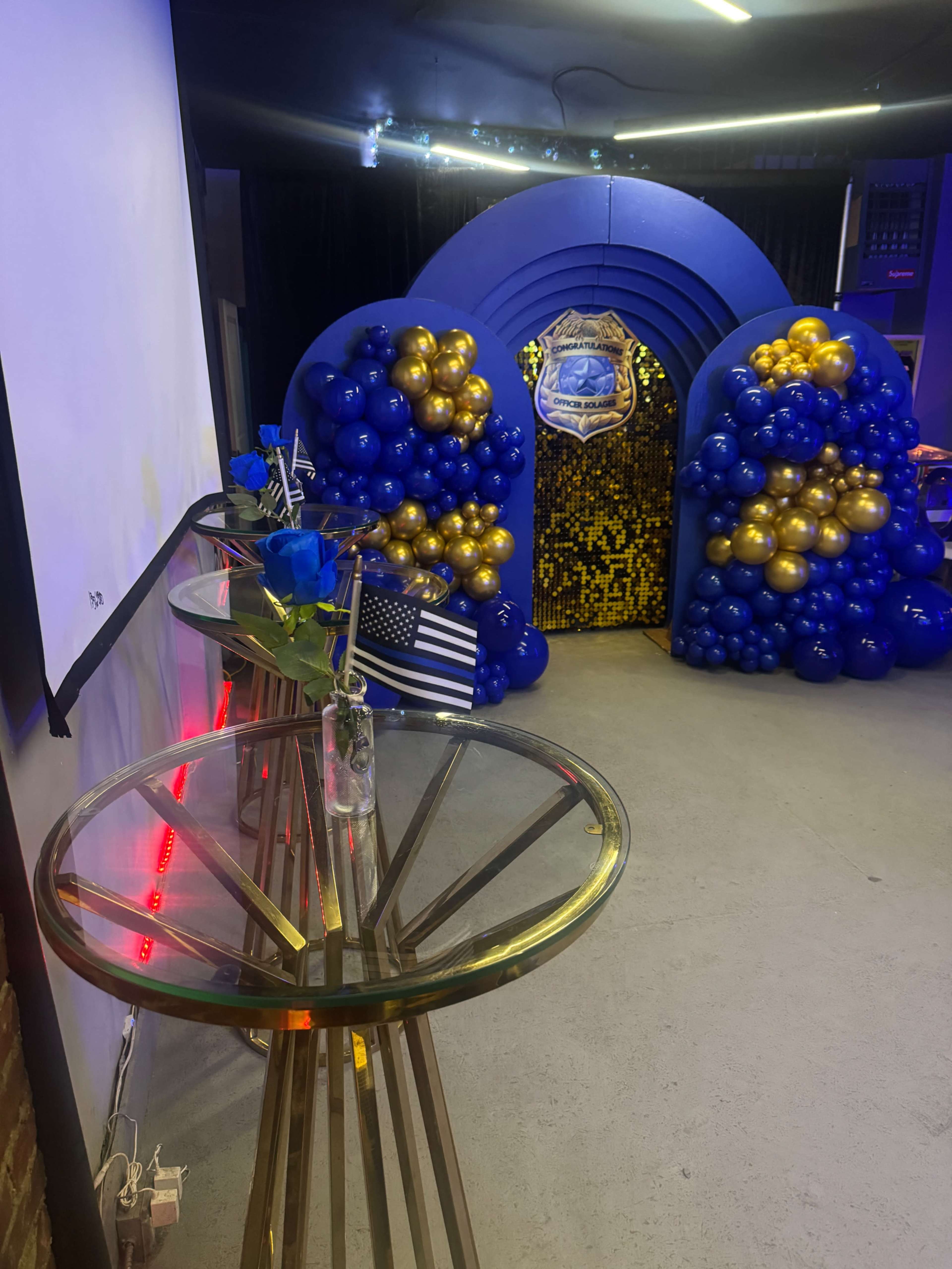The image shows a decorated entrance with a blue and gold balloon arch, leading into a space adorned with a large emblem, and a glass table with a vase and American flag nearby.