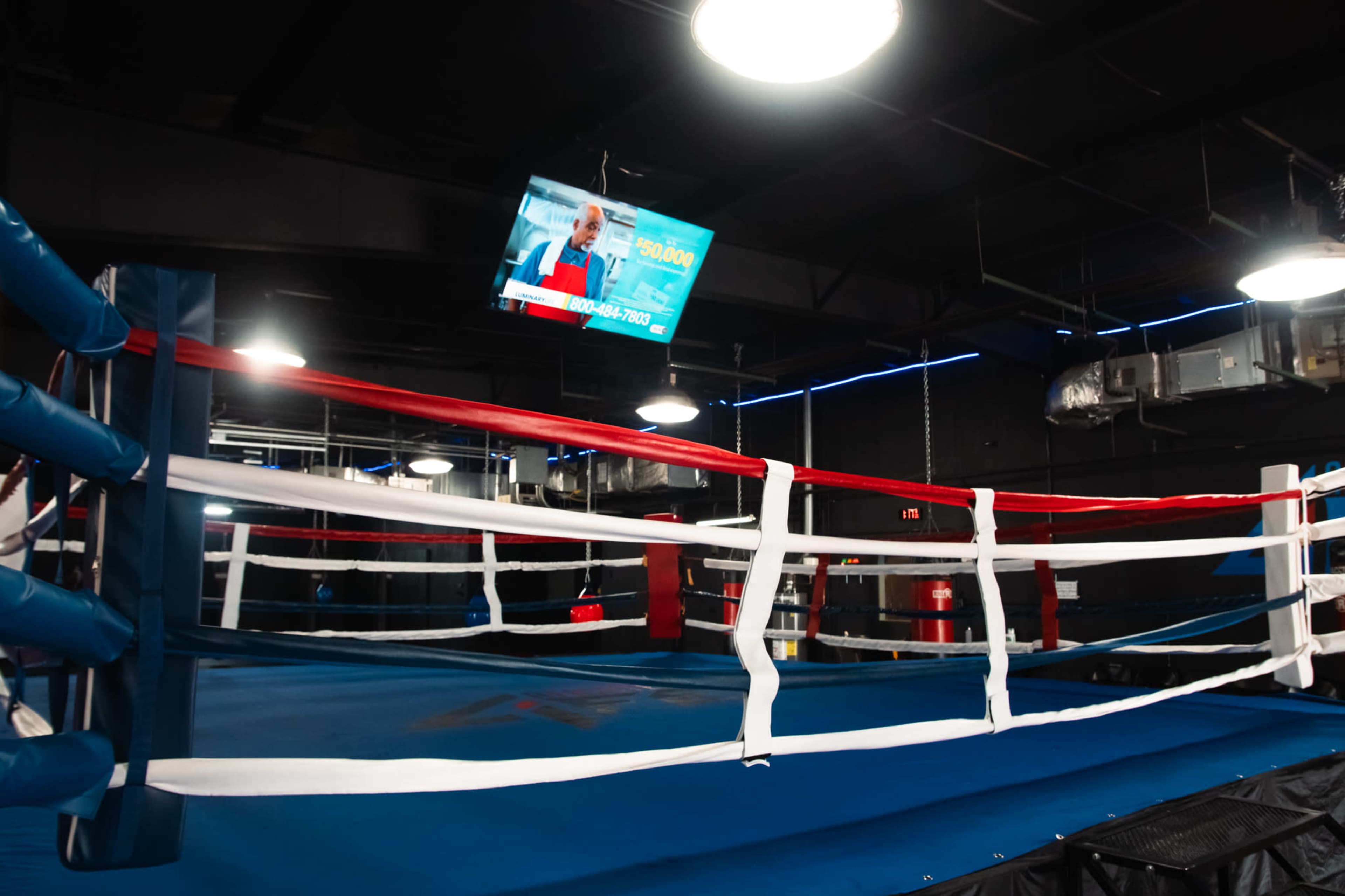 The image shows an empty boxing ring with blue flooring and colored ropes inside a gym, illuminated by overhead lights and featuring a wall-mounted screen.