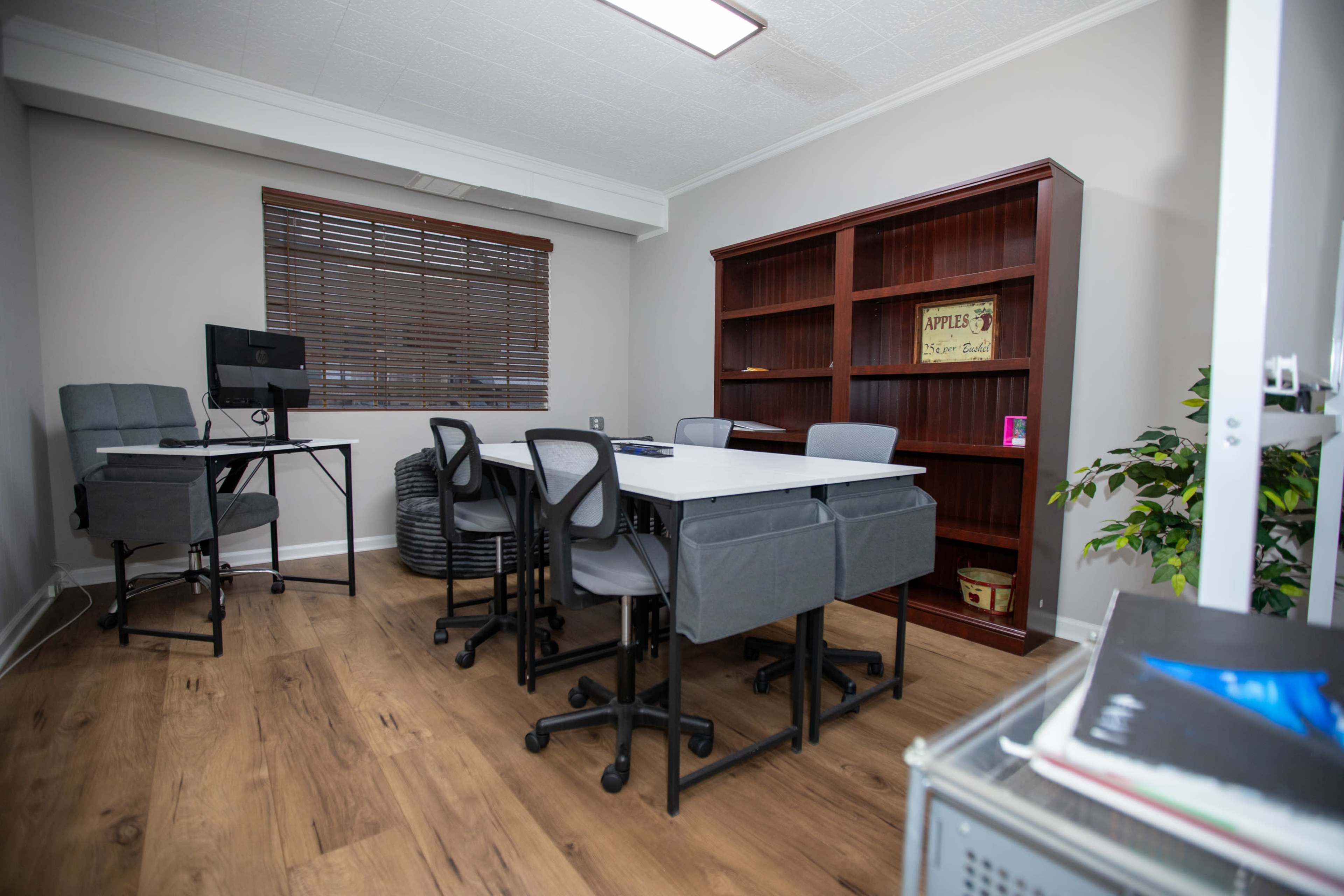 A small office space features a central meeting table surrounded by rolling chairs, a computer station, and a bookshelf along the wall.