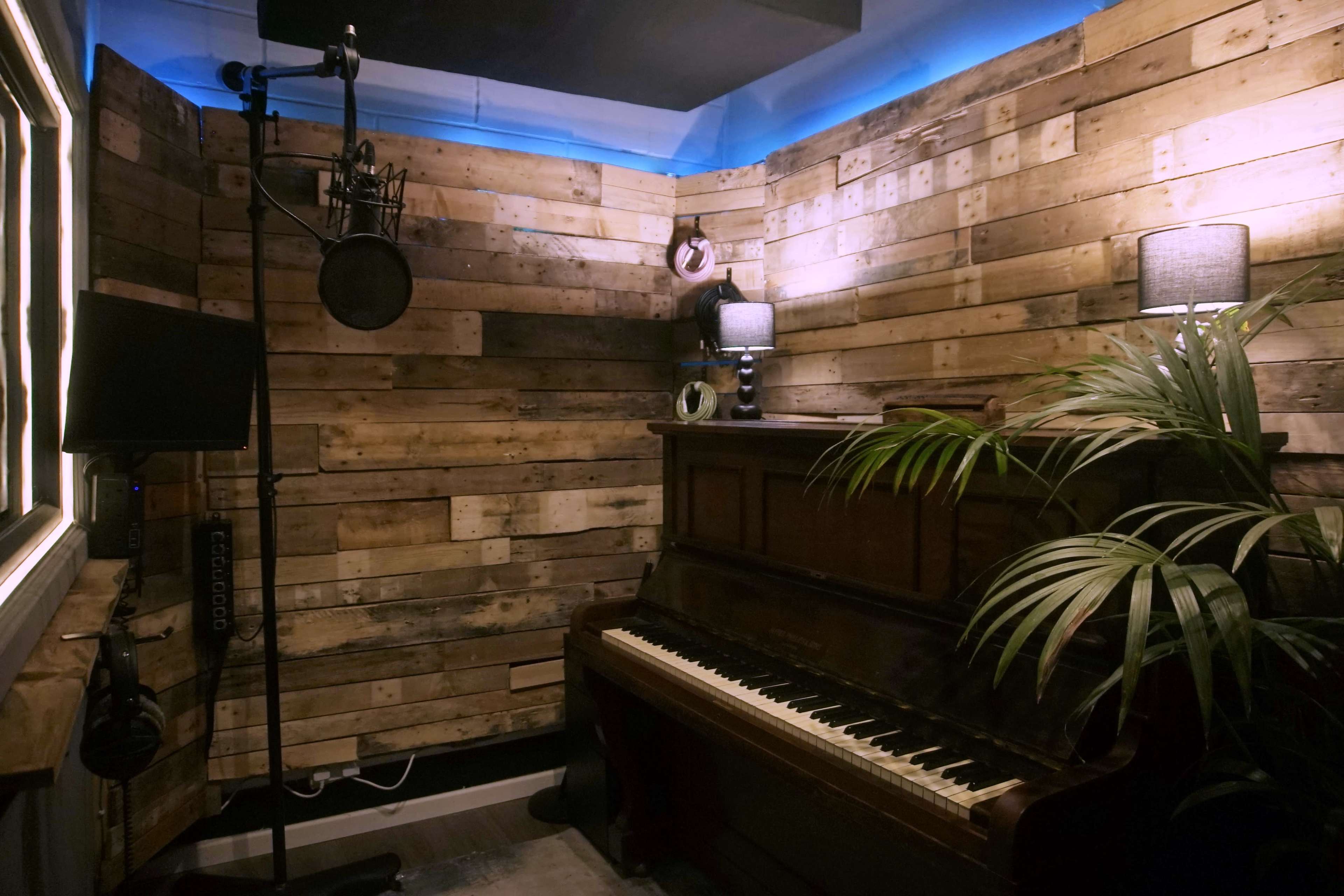 The image shows a small music recording studio with wooden walls, a vintage piano in the corner, and a microphone setup.