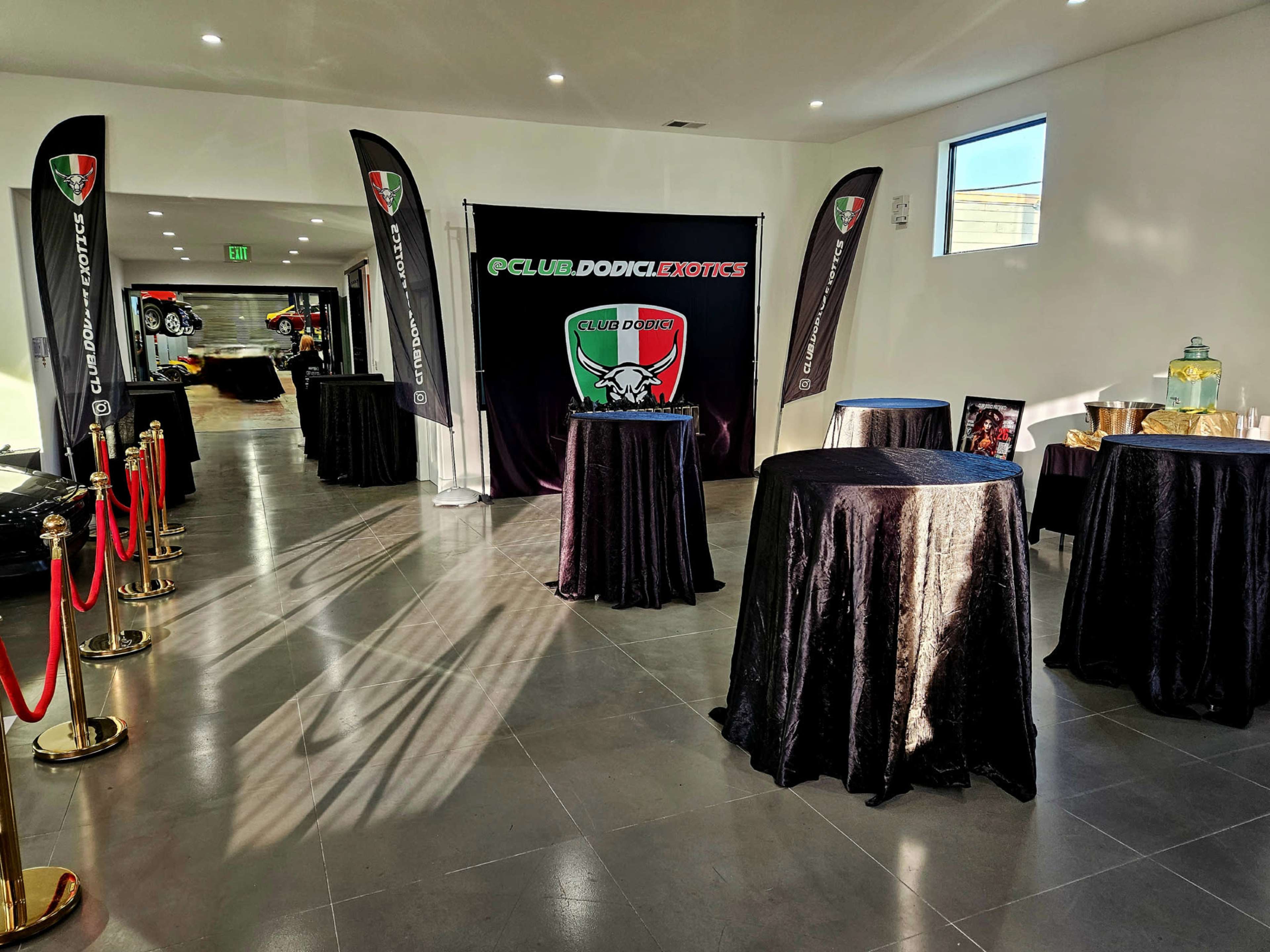 The image shows a spacious indoor event venue with black table covers, red velvet ropes, and banners for "Club Dodge Exotics" displayed on the walls.