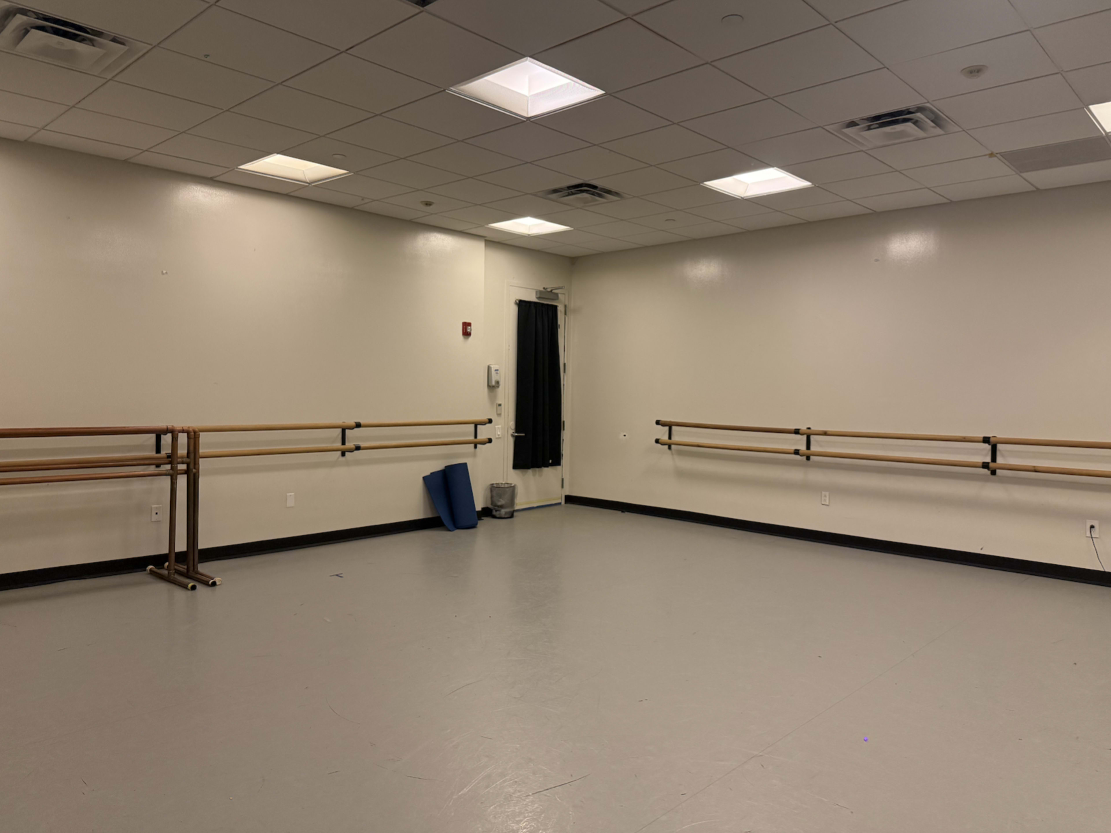 The image shows a spacious dance studio with wooden ballet bars attached to the walls and gray flooring.