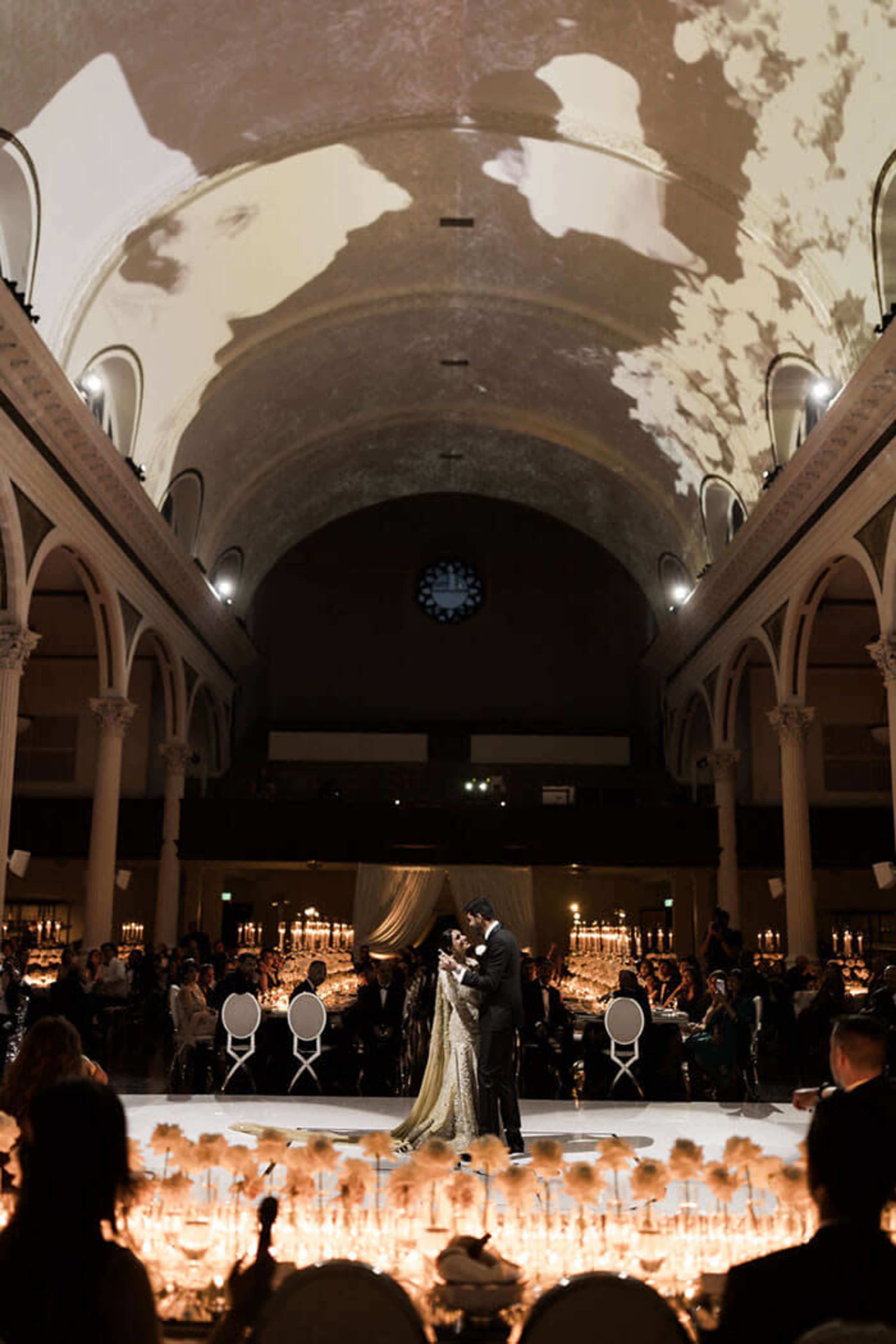 A couple is exchanging vows at a centrally located altar in a grand hall adorned with elegant lighting and an audience seated around the perimeter.