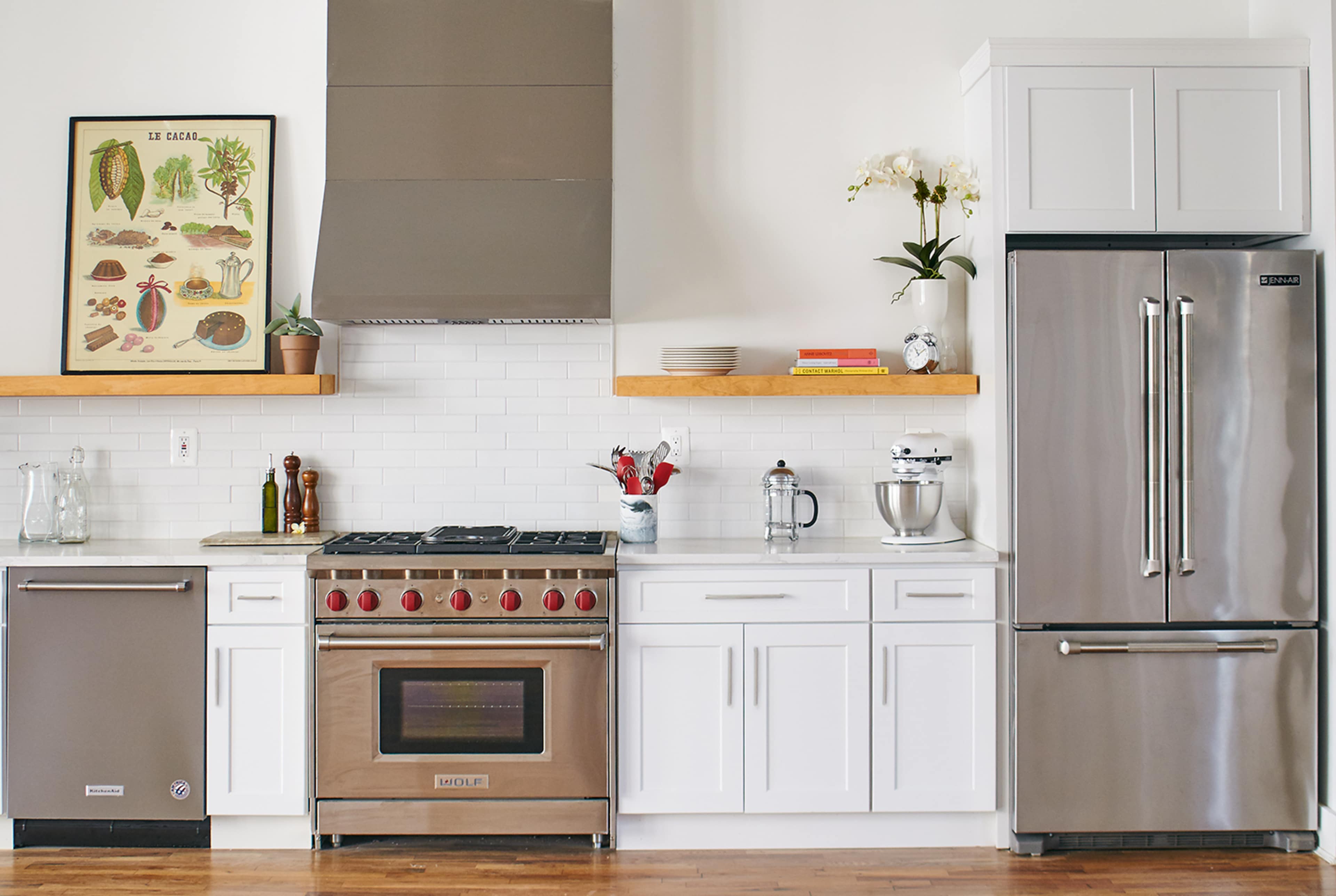 A modern kitchen with stainless steel appliances, white cabinetry, and wooden shelves displaying a poster and kitchen items.