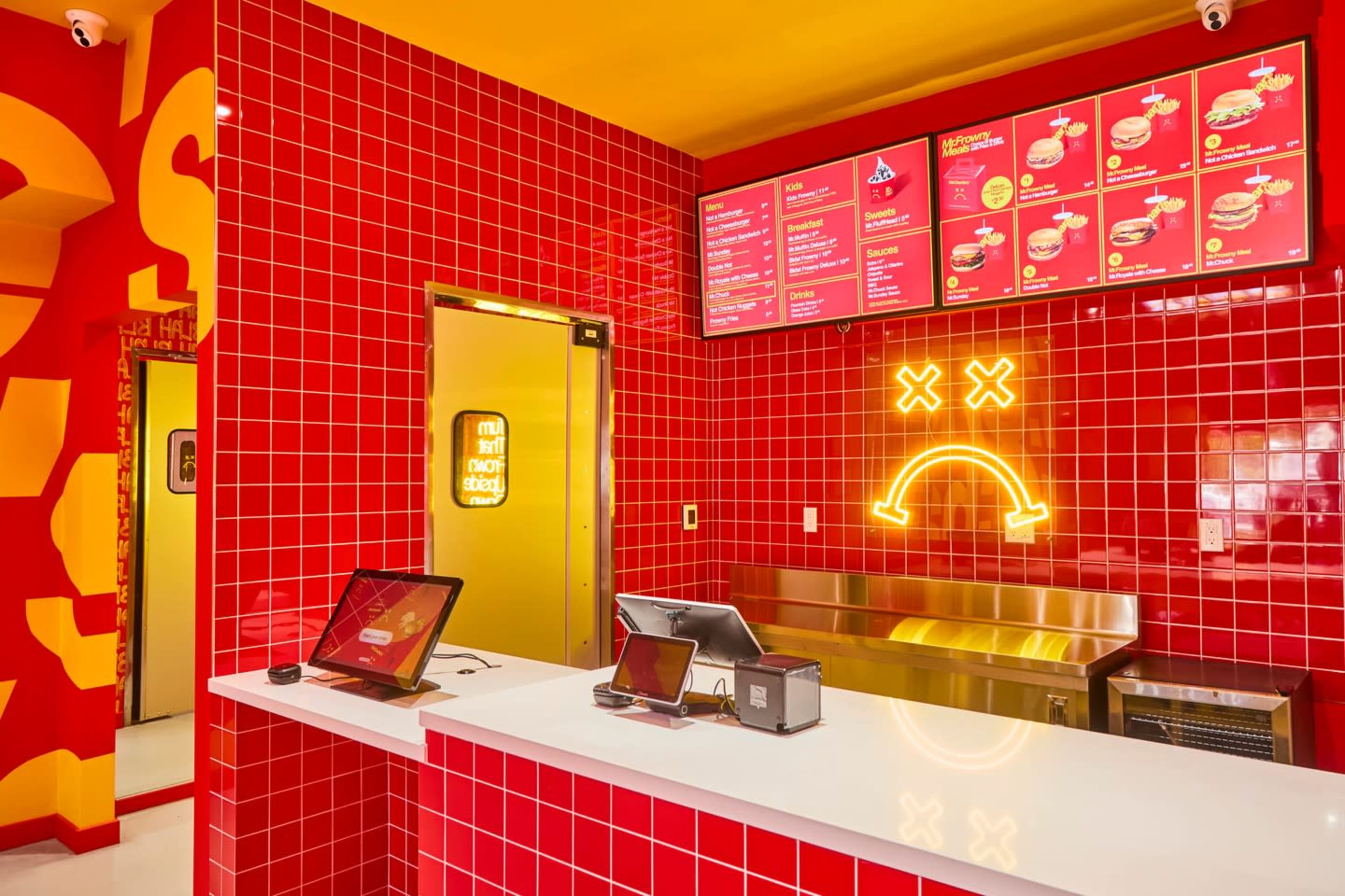 The interior of a fast-food restaurant features bright red tiled walls, a menu board with colorful items, and a smiling neon sign above a service counter.