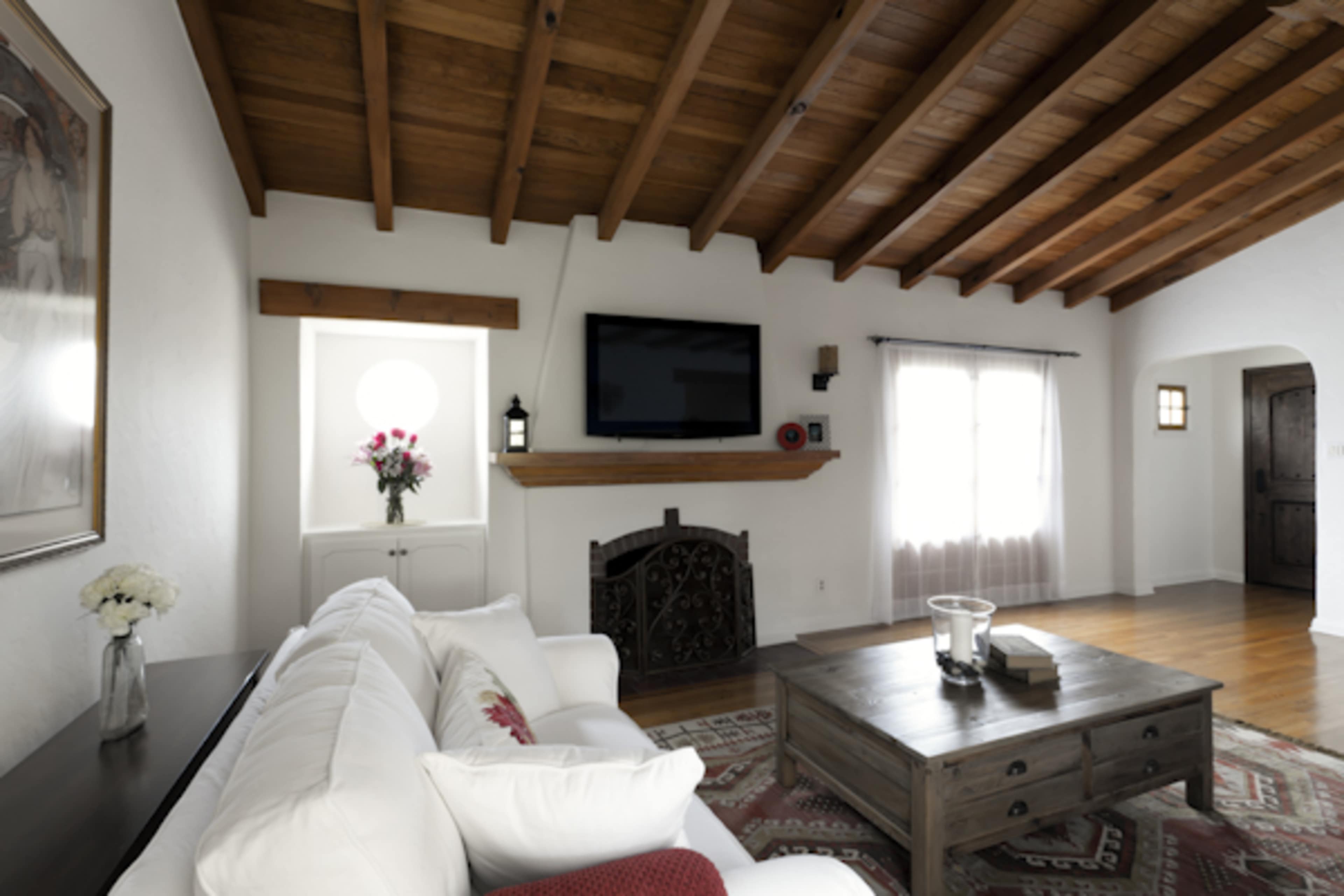 The image shows a living room with a white sofa, a wooden coffee table, a flat-screen TV mounted above a wooden mantel, and large windows with sheer curtains.