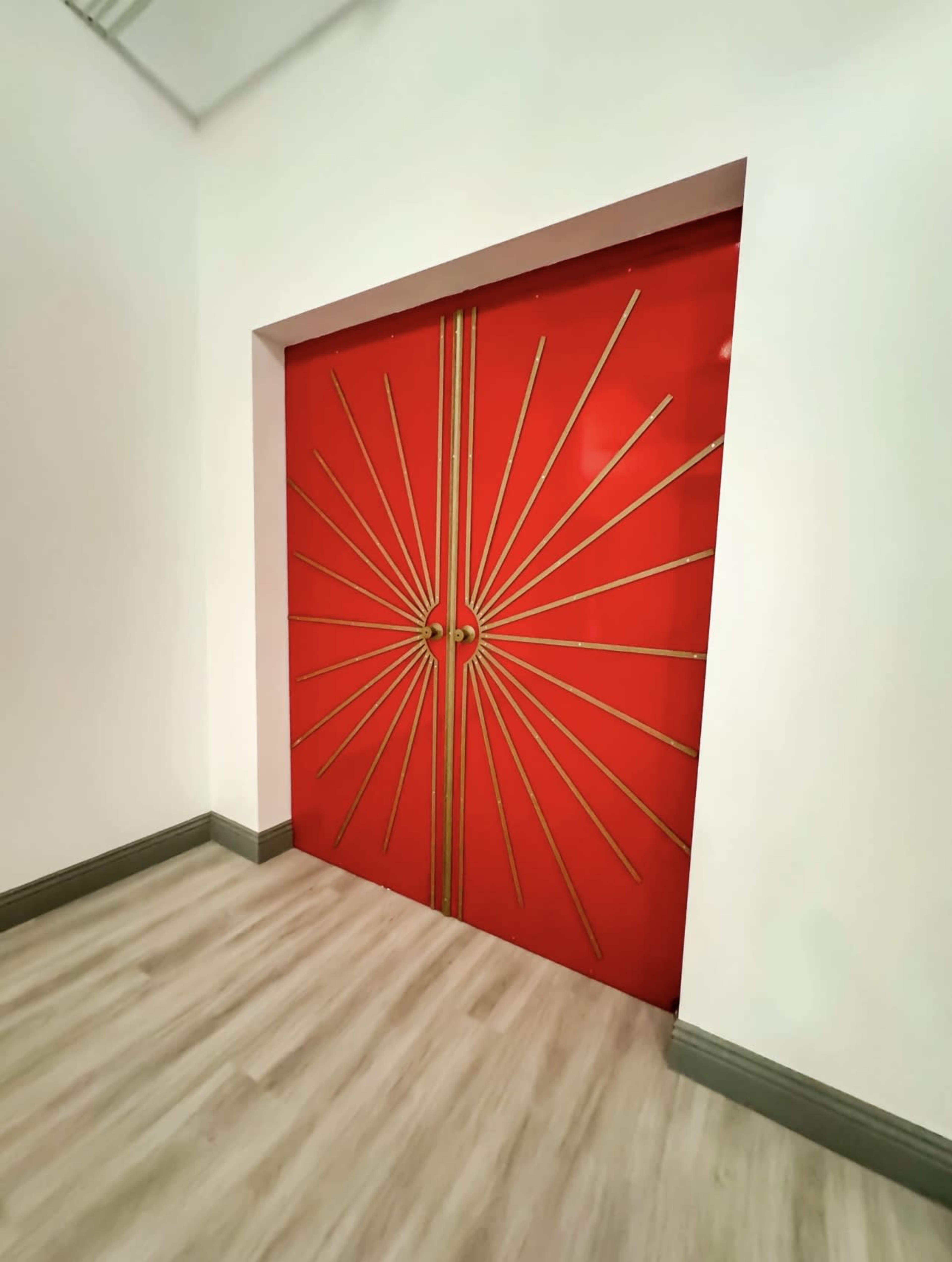 A red double door with a sunburst design and golden accents, set against a light-colored wall and wooden floor.