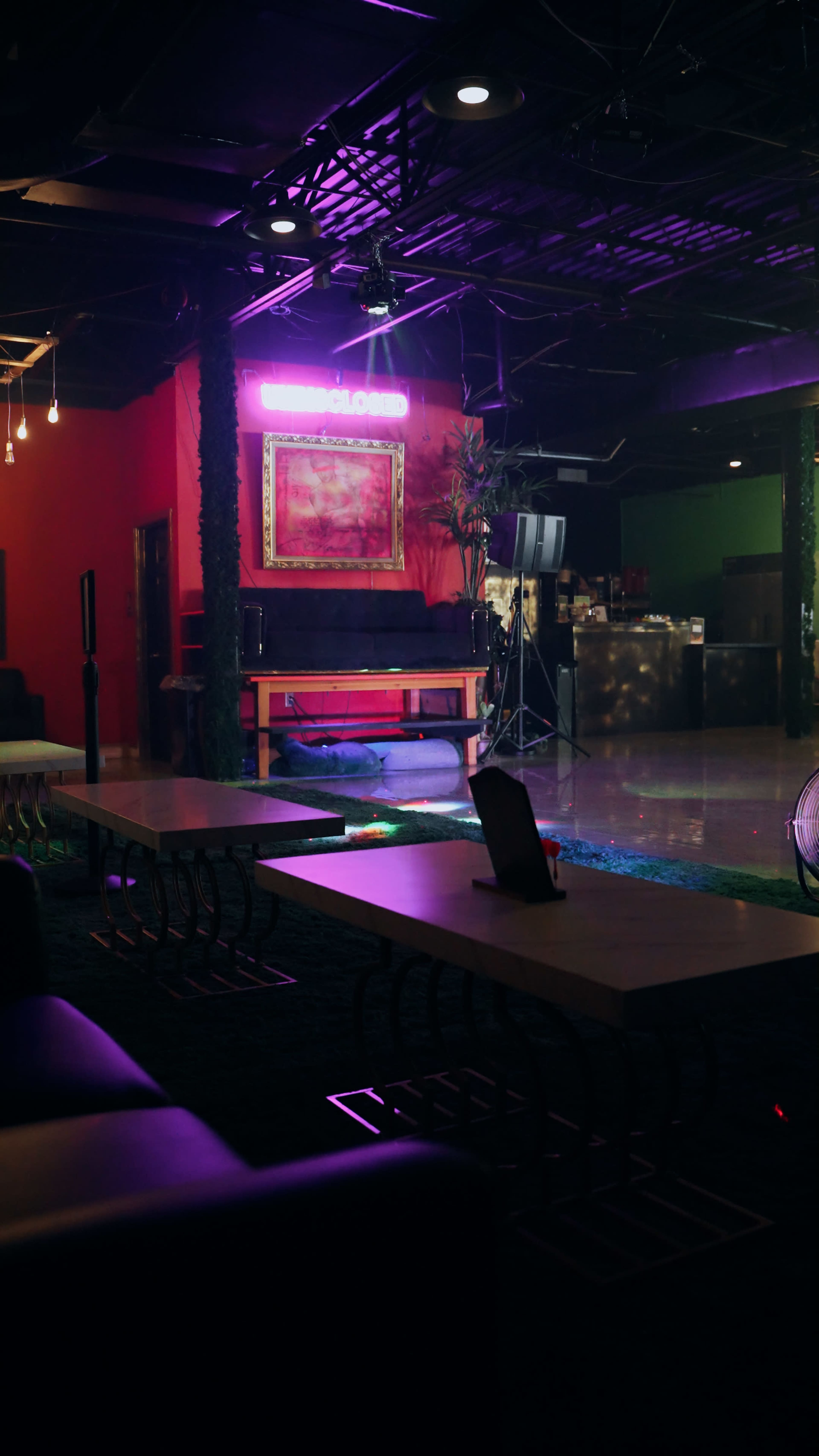 The image shows a dimly lit lounge area with a bar, a neon sign, and several tables arranged on a carpeted floor.