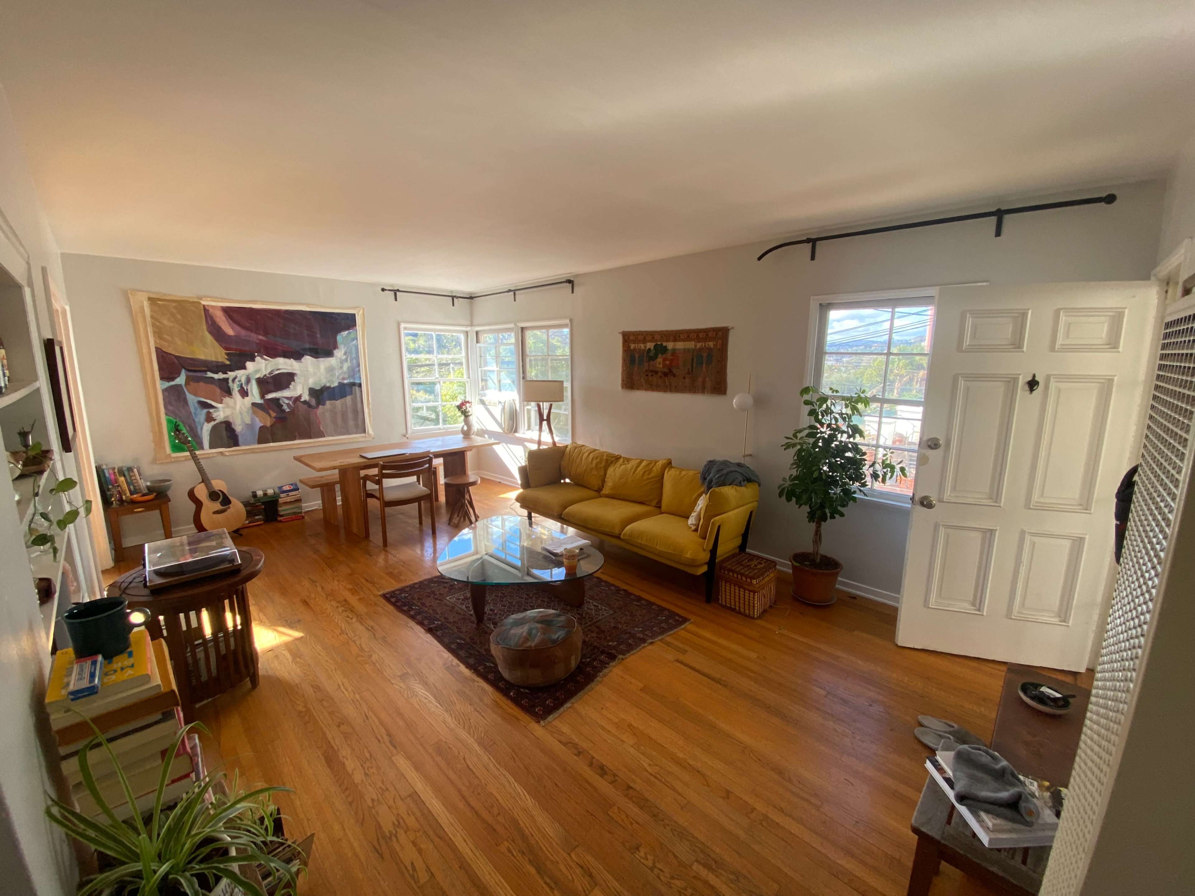 A spacious living room features a yellow sofa, a glass coffee table, a dining area with a wooden table, and large windows letting in natural light.