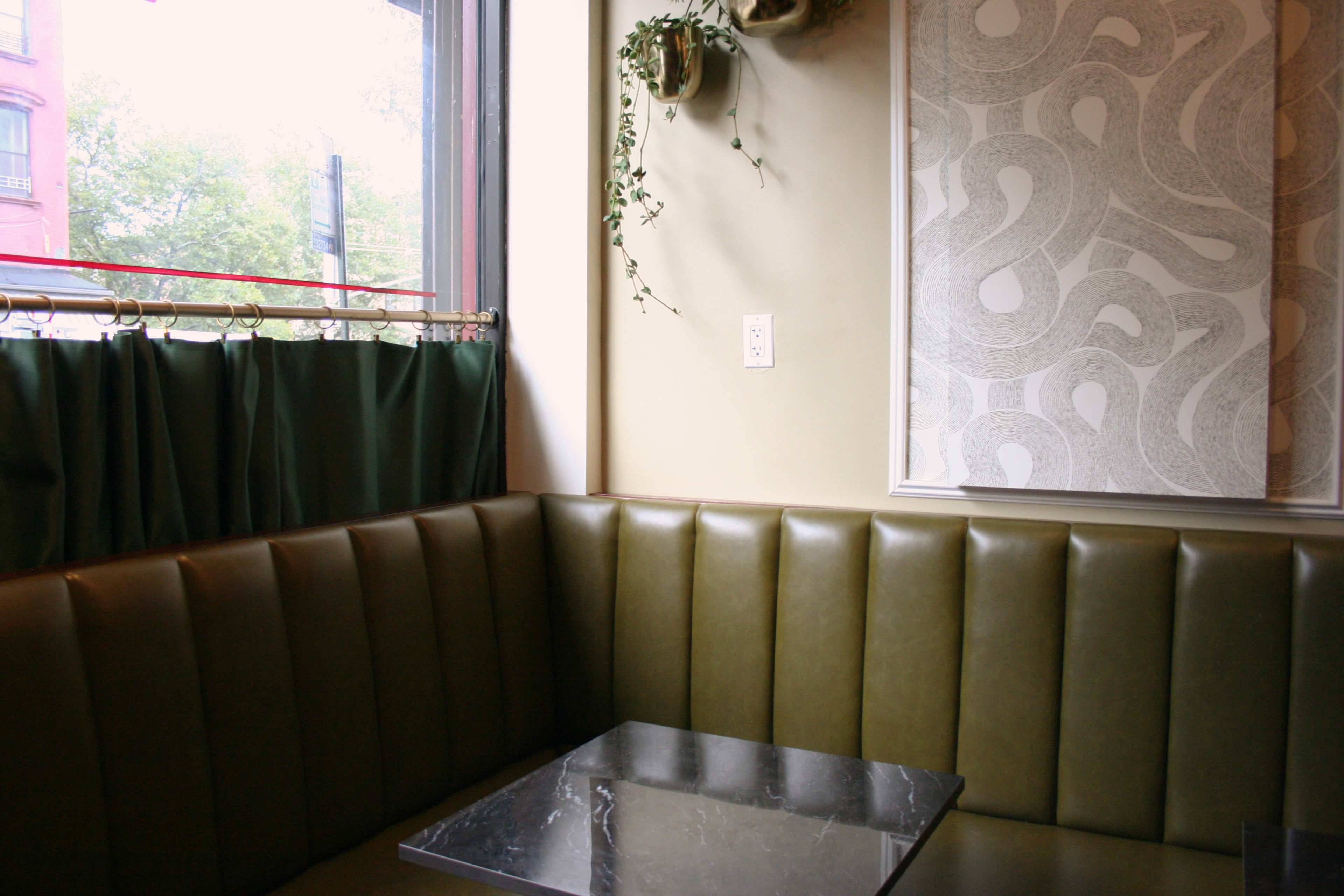 The image shows a cozy corner of a café featuring a green bench seat, a marble-top table, and a window draped with dark curtains, complemented by decorative wall elements.