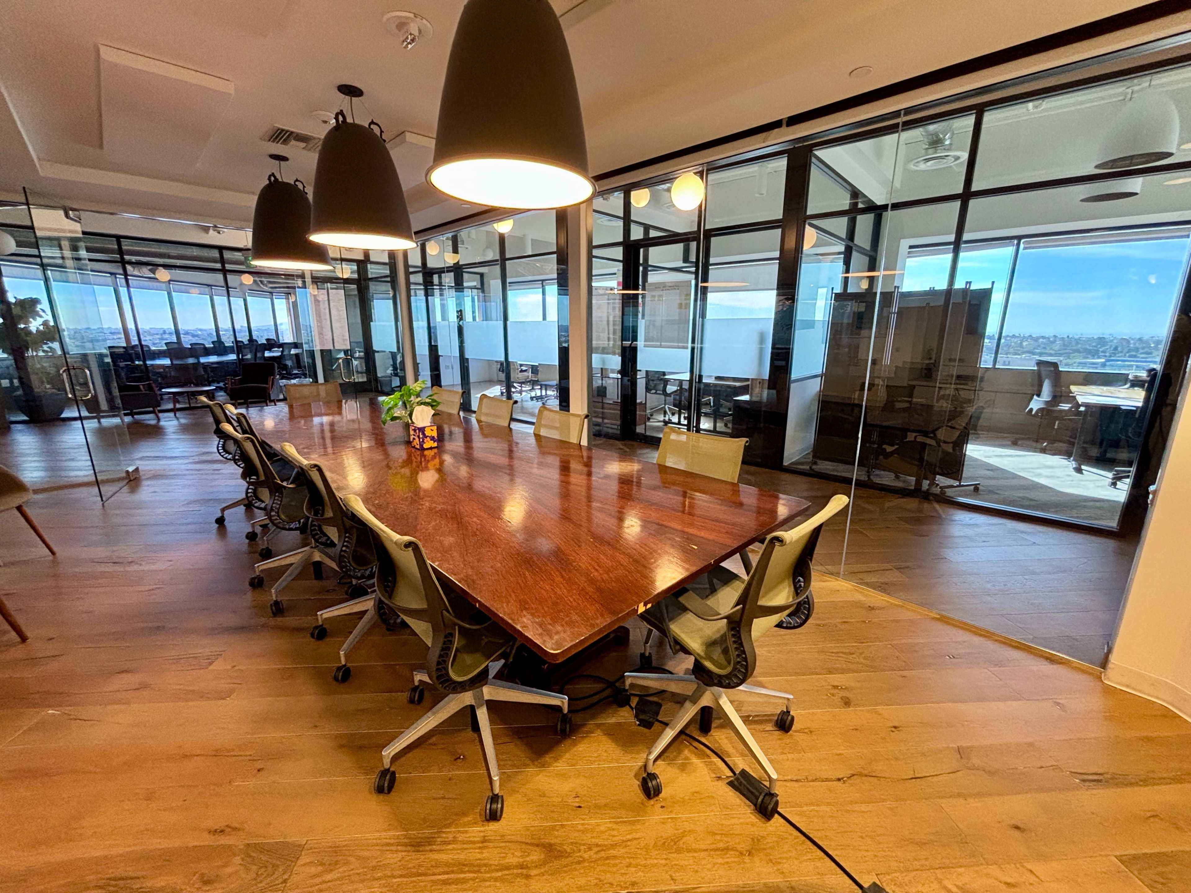 A long wooden conference table is surrounded by office chairs in a glass-walled meeting room with large windows overlooking a cityscape.