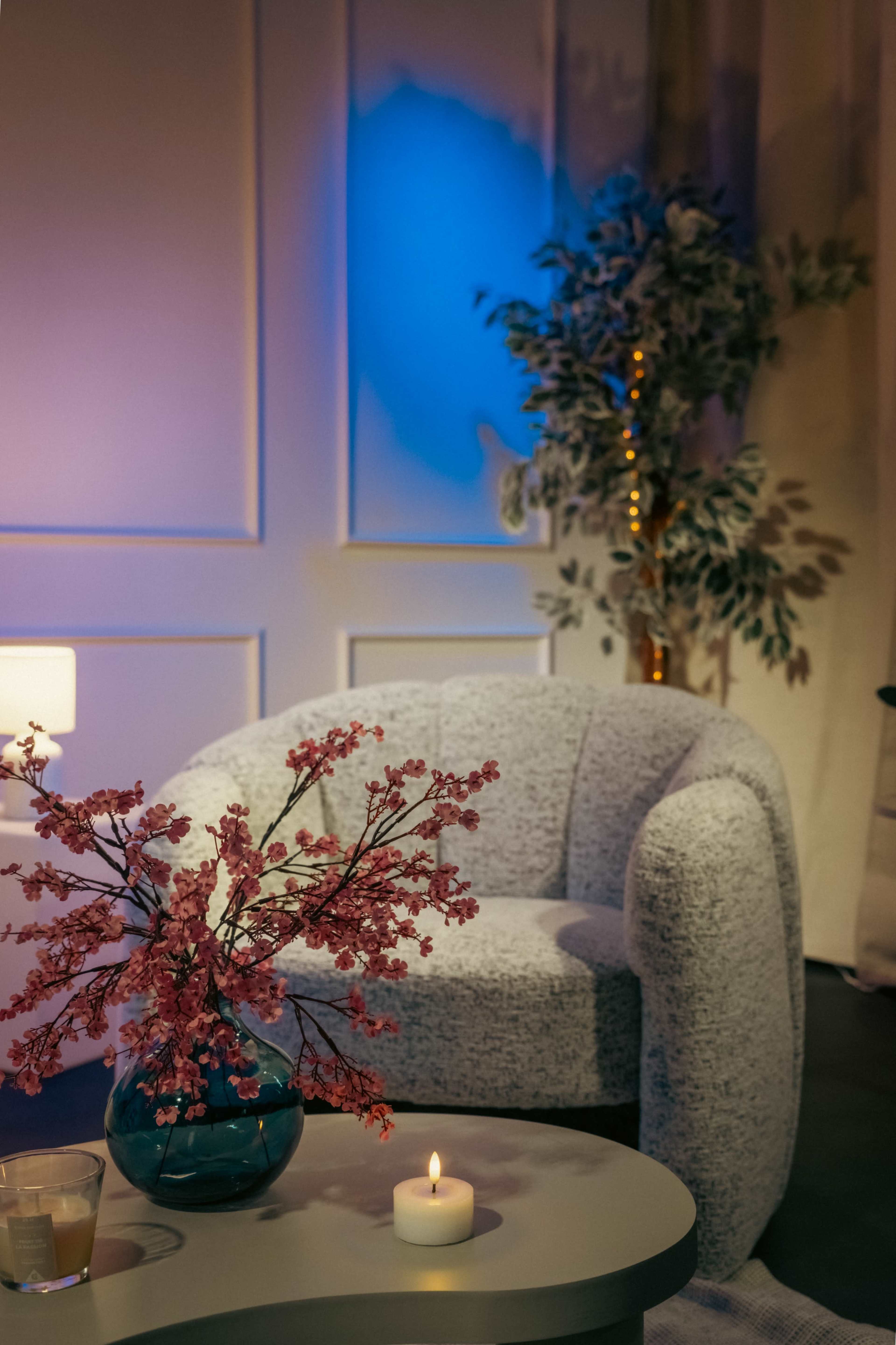 A small, cozy living space features a textured gray chair, a flower arrangement in a blue vase, and a lit candle on a coffee table, with a decorative plant in the background.