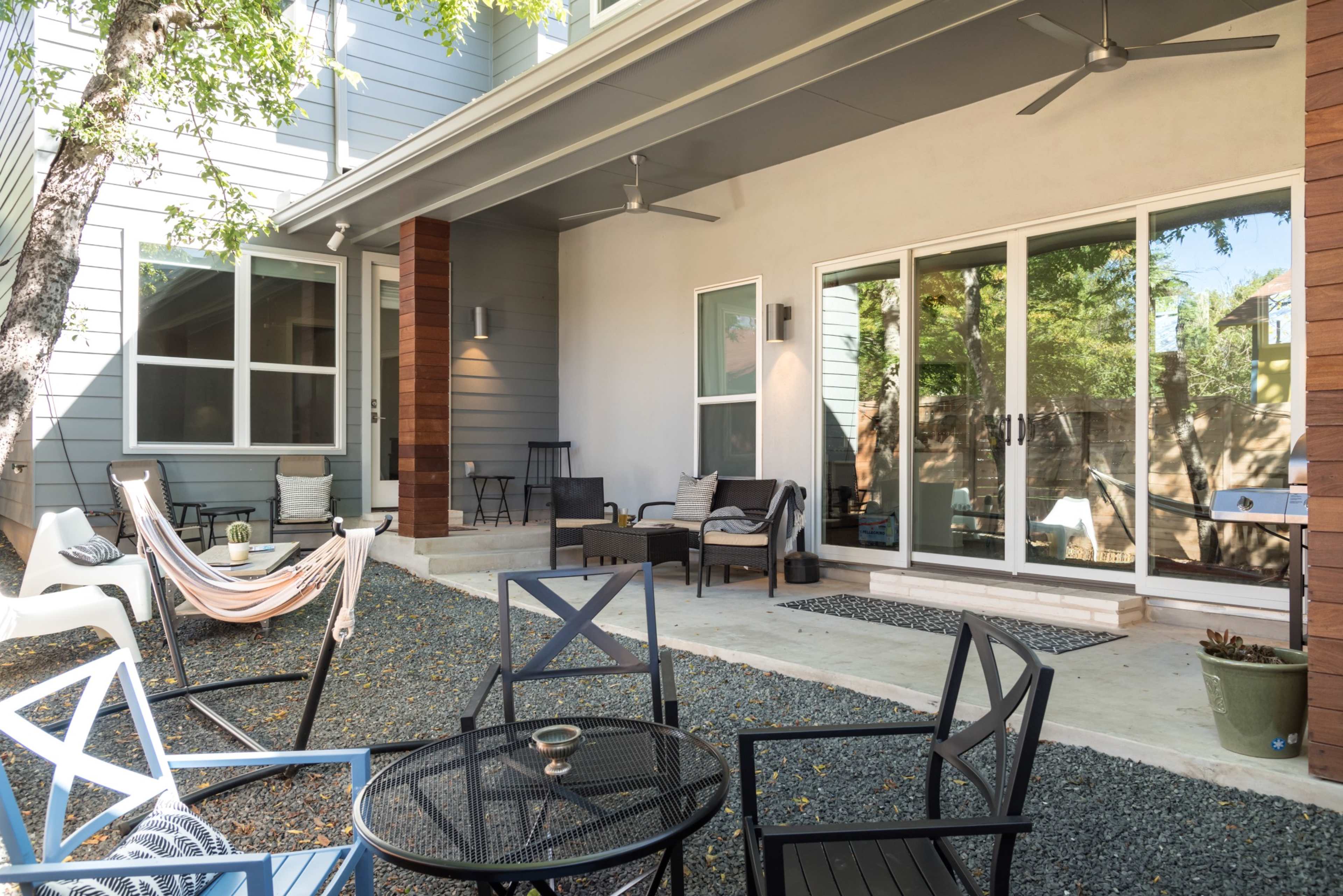 A patio area with outdoor seating, a hammock, and gravel flooring, surrounded by trees and sliding glass doors leading inside.