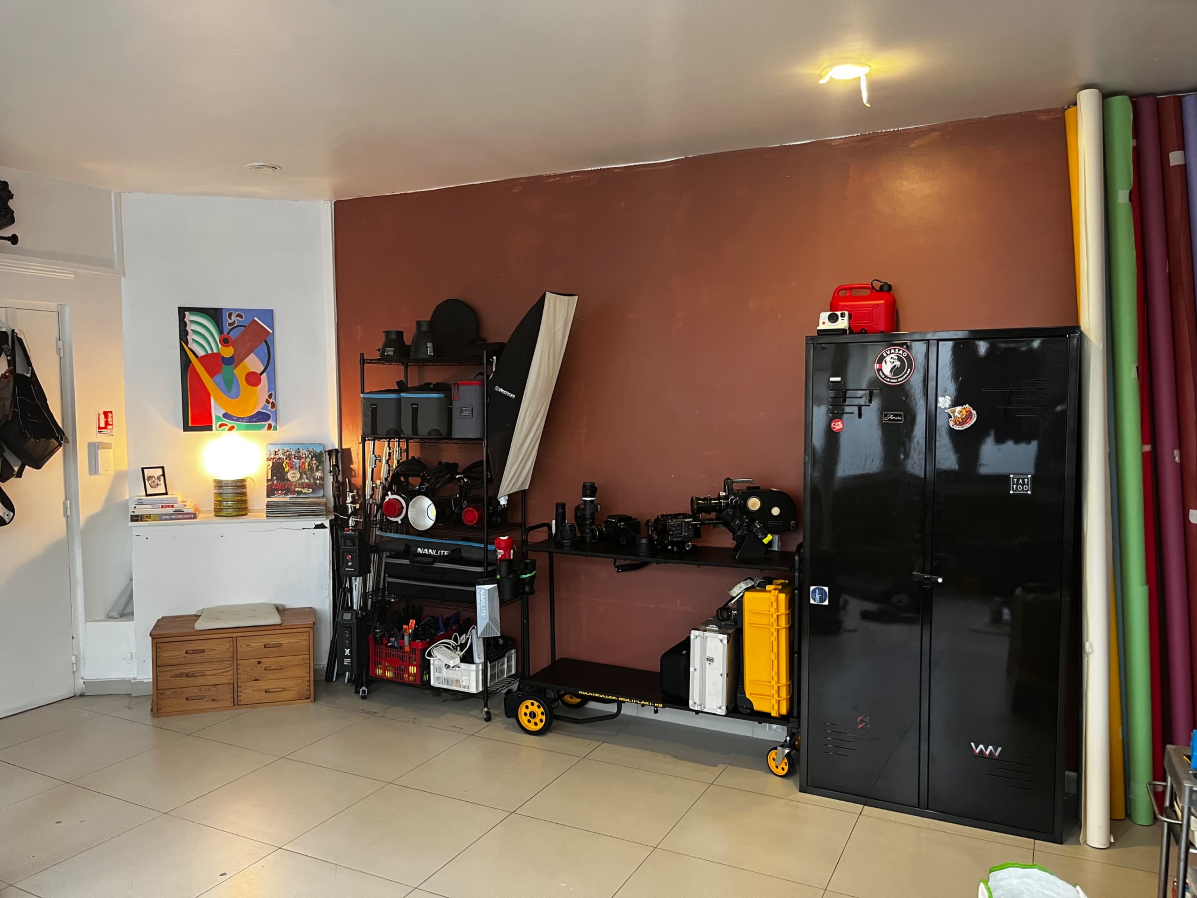 The image shows a room featuring a brown wall, a black cabinet, a storage cart with various tools, a small wooden storage unit, and several rolls of paper in different colors.