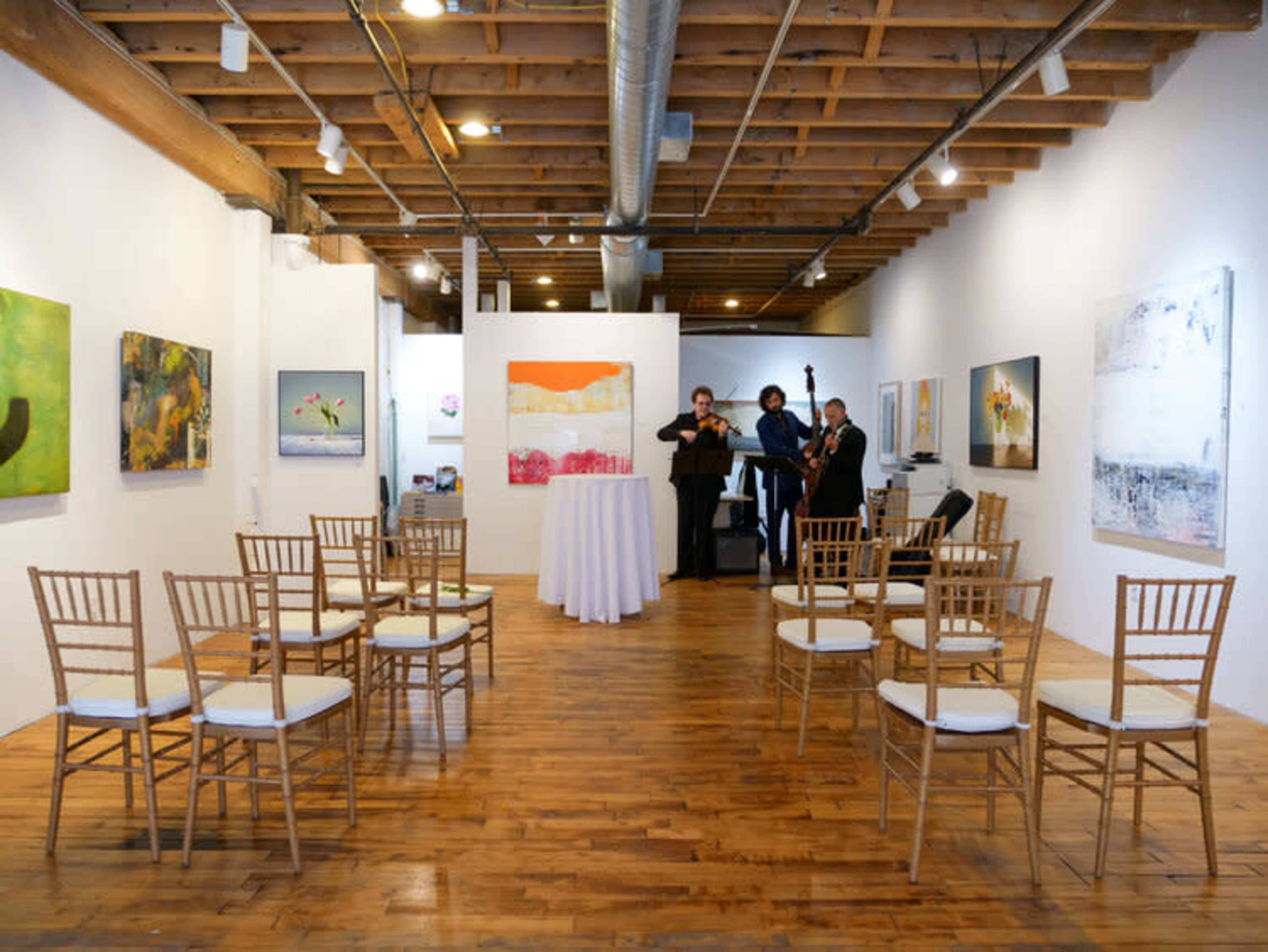 A spacious art gallery features wooden flooring and several chairs arranged in two semi-circles, with musicians performing near colorful abstract paintings.