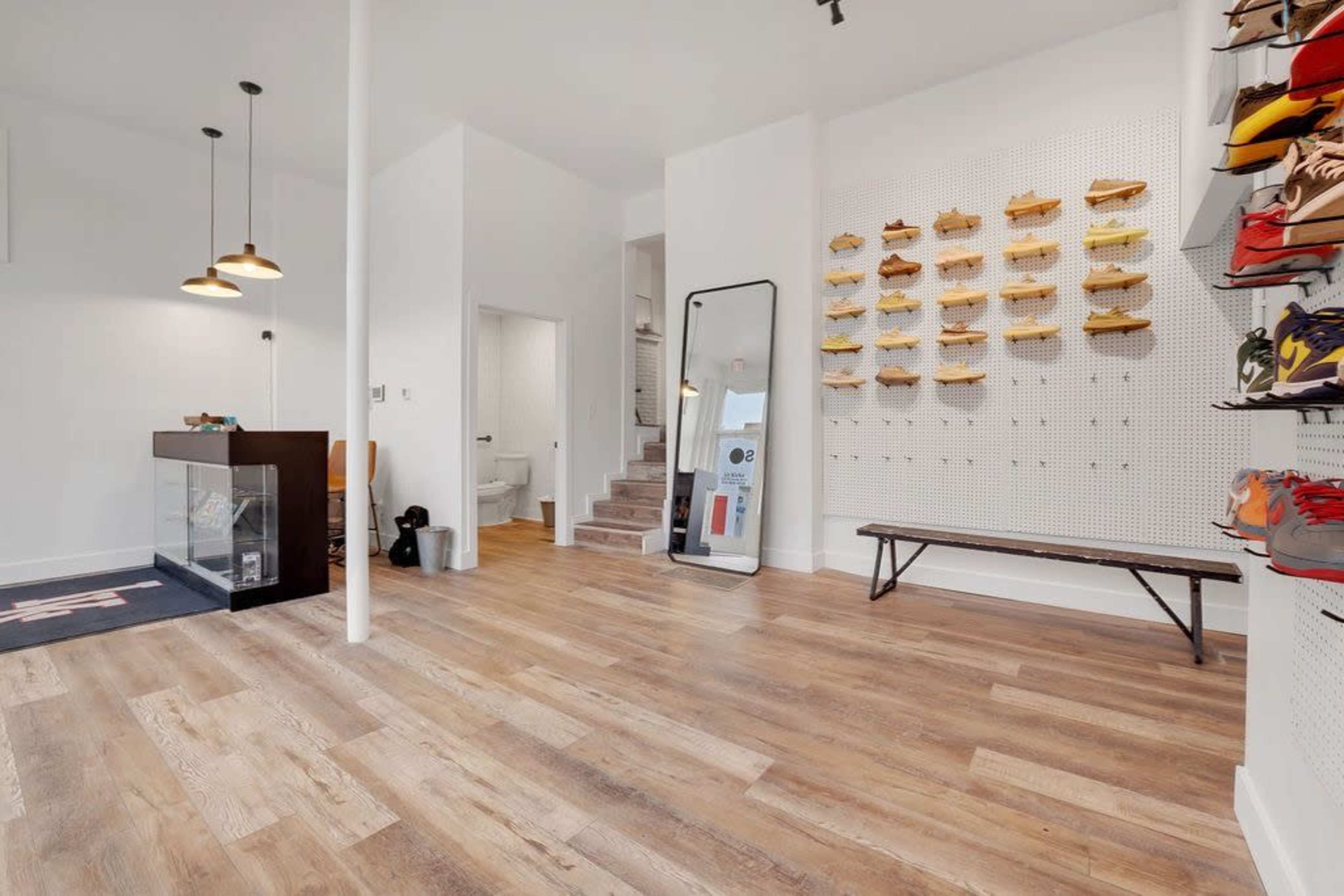 The image shows a modern retail space with wooden flooring, a display wall of shoes, a mirror, and a minimalist counter area.