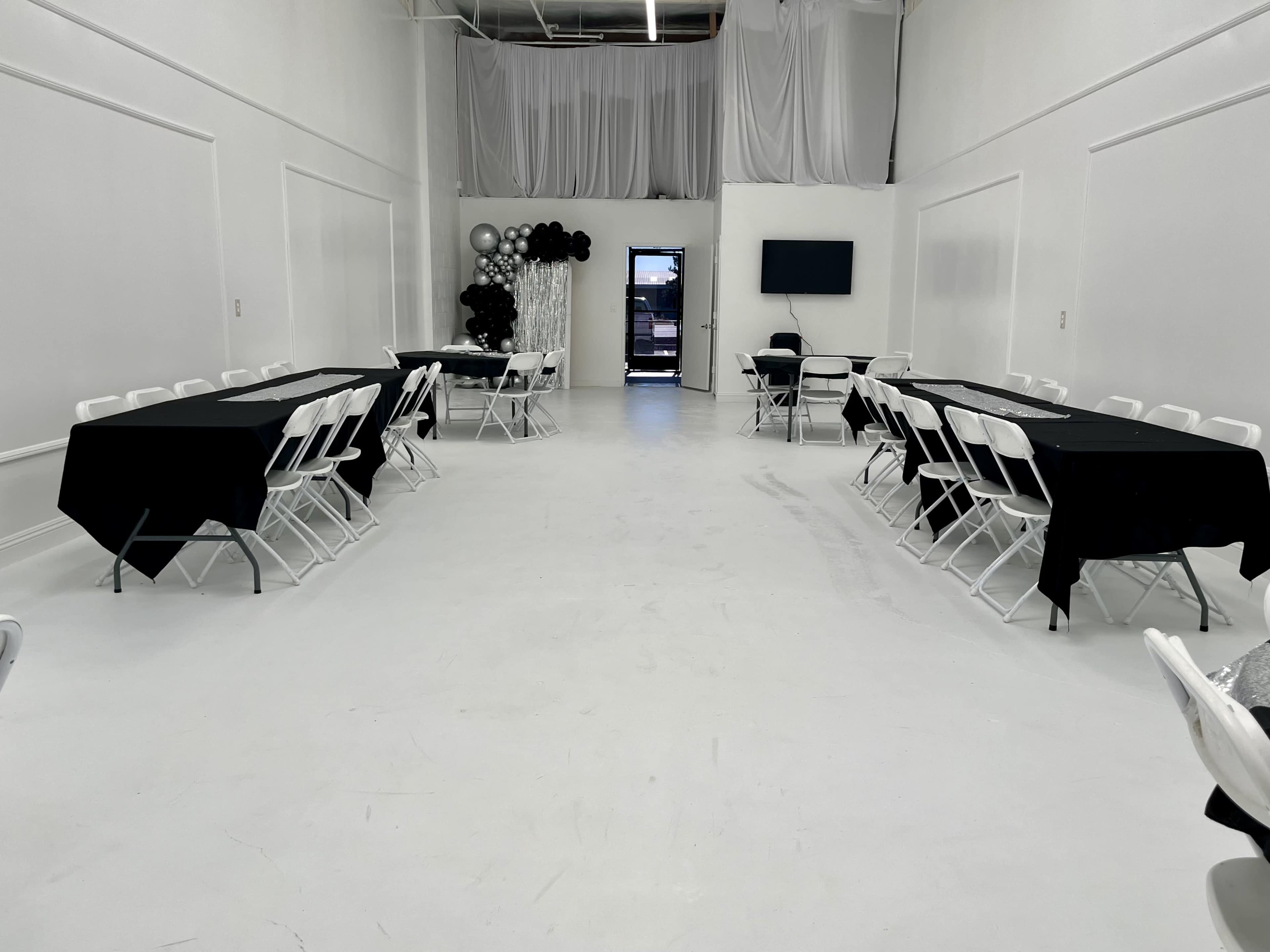 The image shows a spacious, empty room set up with black tablecloths over white folding tables arranged in two rows, with a black and white decorative arrangement in the corner.