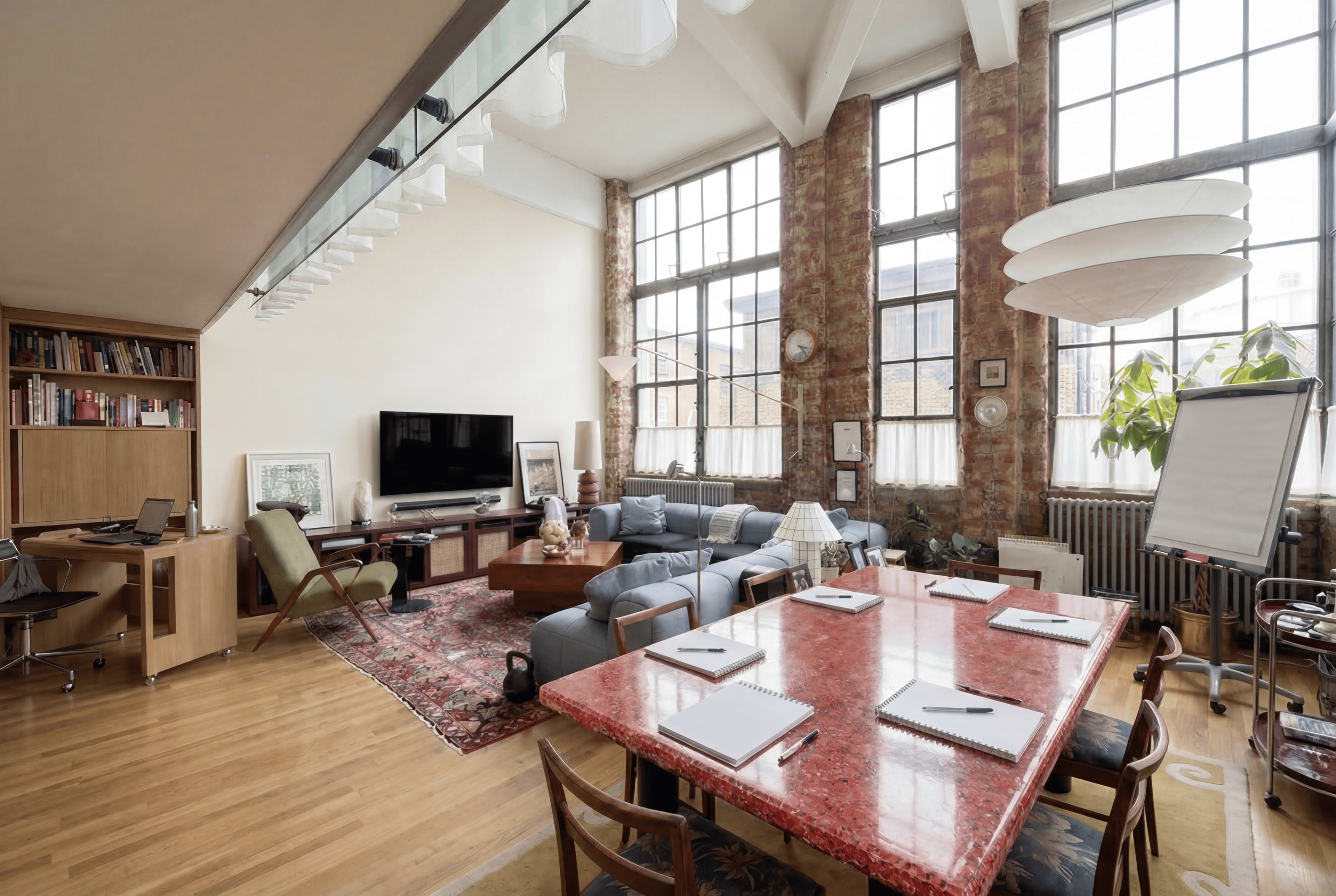 The image shows a spacious, modern loft-style office with large windows, wooden floors, and a mix of seating areas and a long dining table set for a meeting.