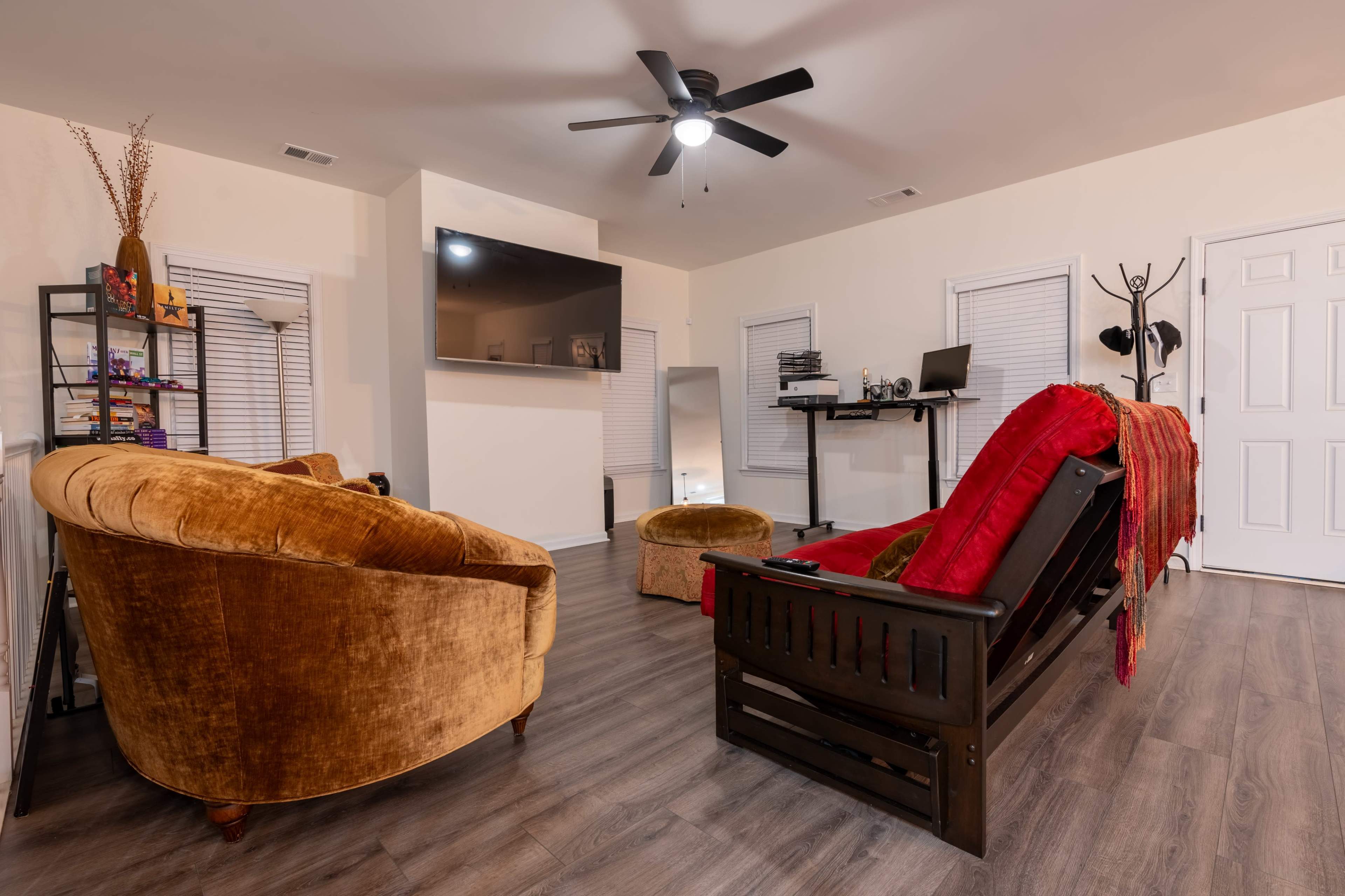 The image shows a cozy living room featuring brown velvet chairs, a wooden bench, a small desk with a computer, and a wall-mounted TV, all set on a wood-patterned floor.