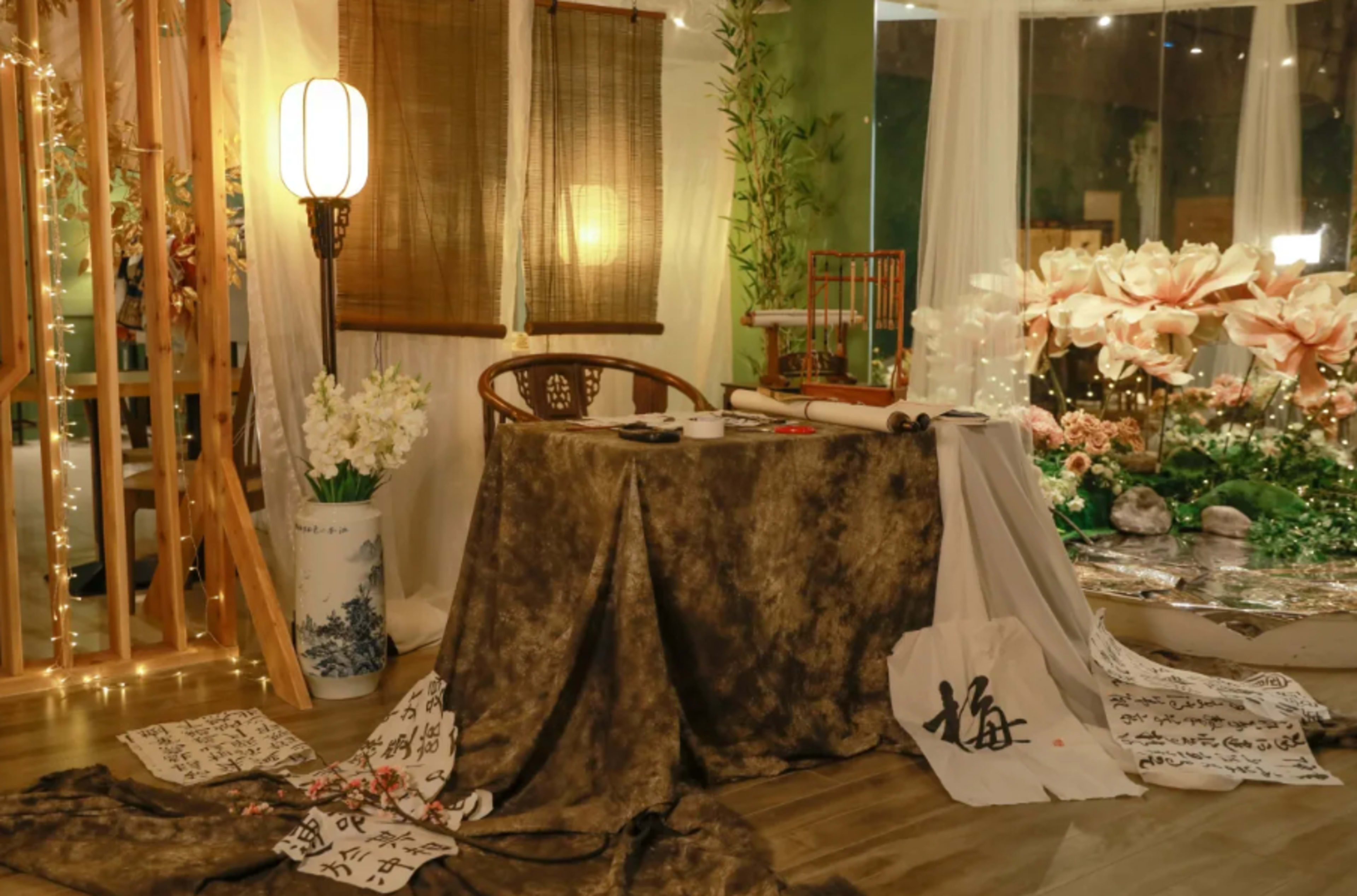 A cozy indoor space features a table covered with a dark cloth, surrounded by scrolls of calligraphy and decorative elements like a floor lamp and plants.