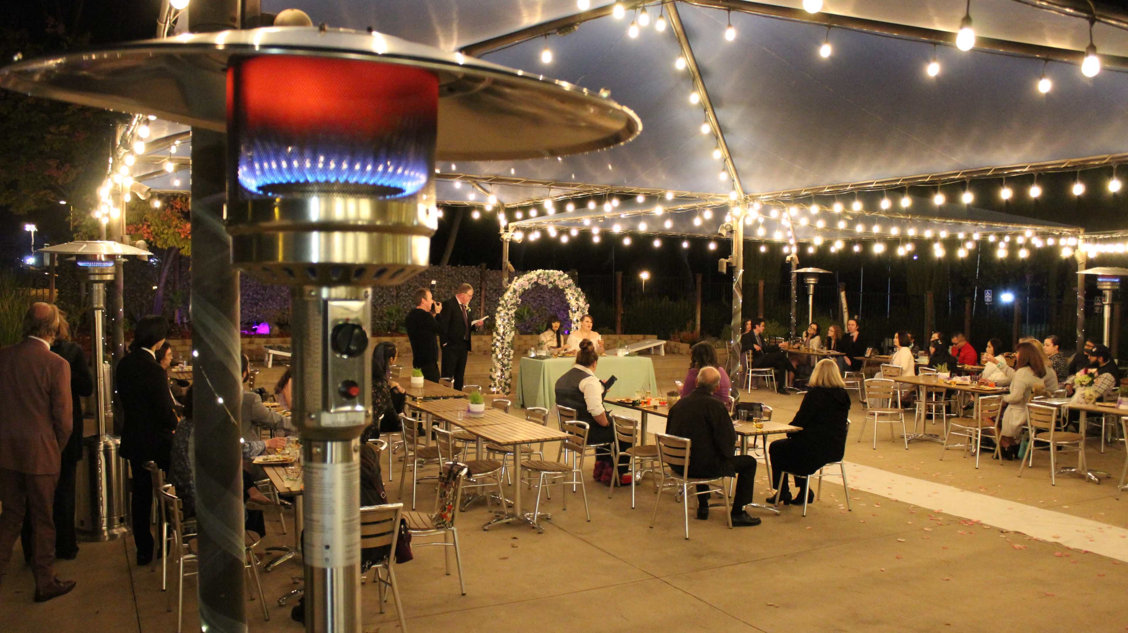 The image shows an outdoor event under a tent with string lights, featuring guests seated at tables and a decorated area at the front.