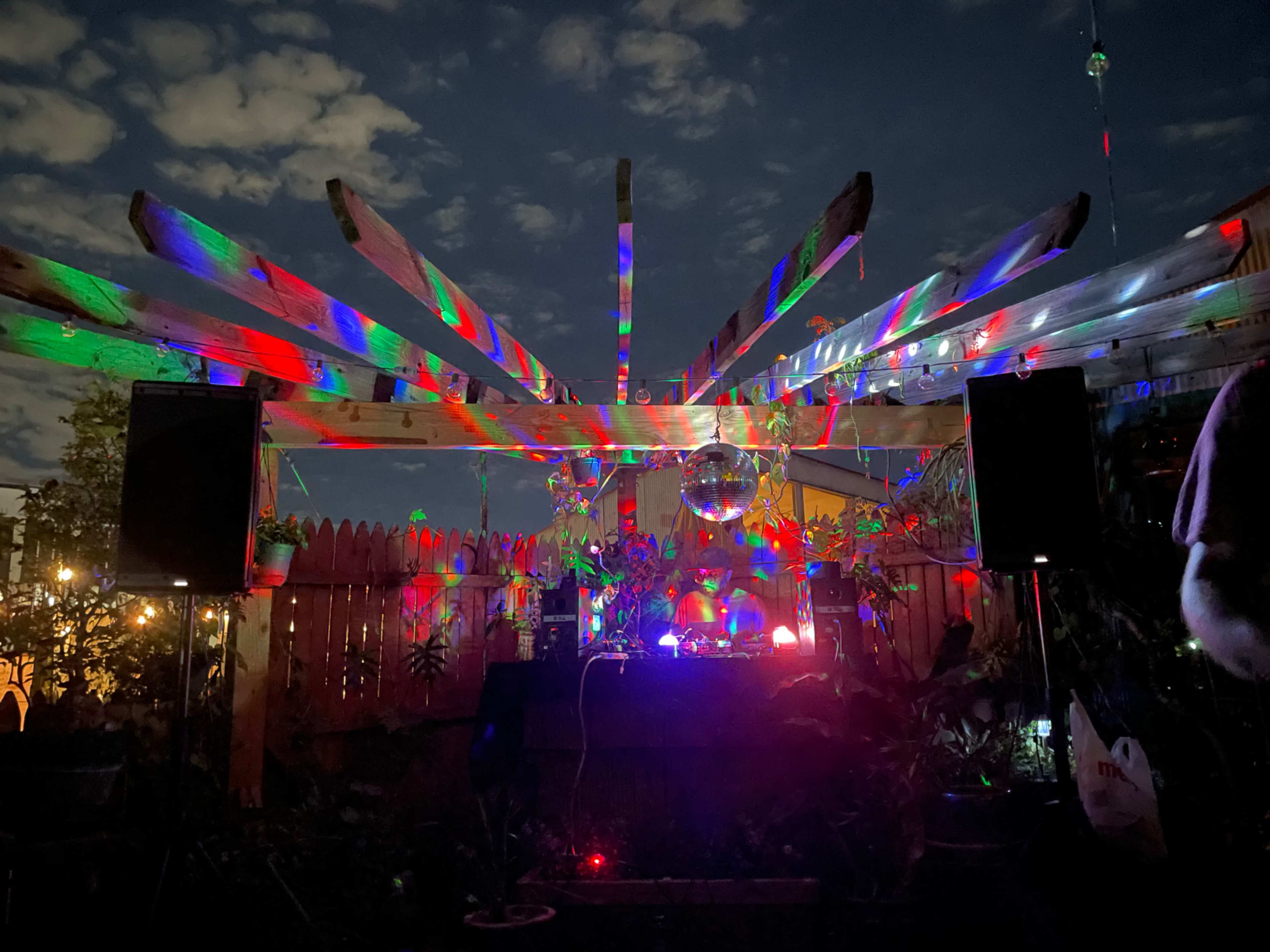 A backyard party setup features a disco ball under a wooden structure, illuminated by colorful lights against a night sky.