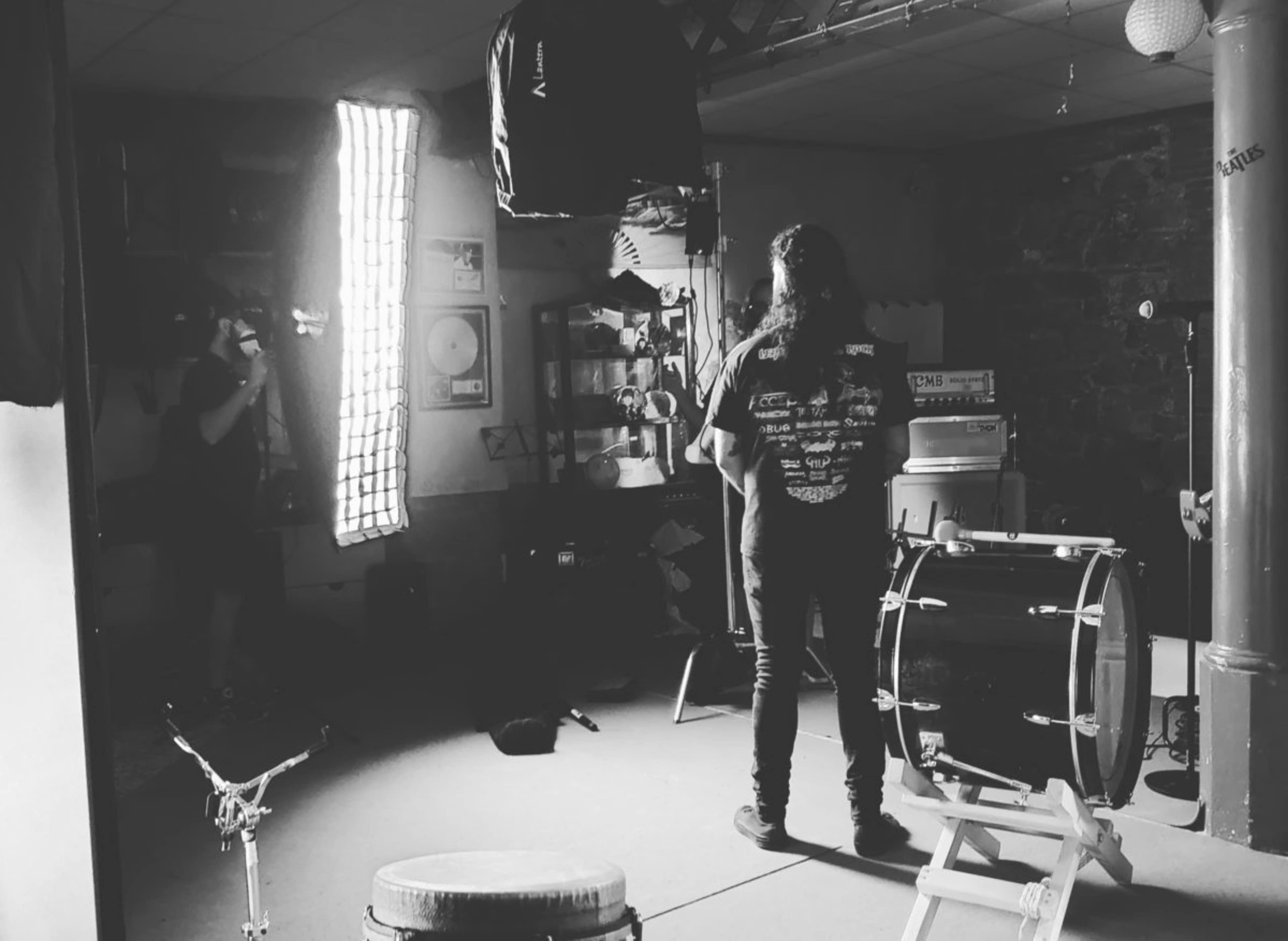 A person stands in a dimly lit room with instruments and music equipment while another individual is taking a photo.