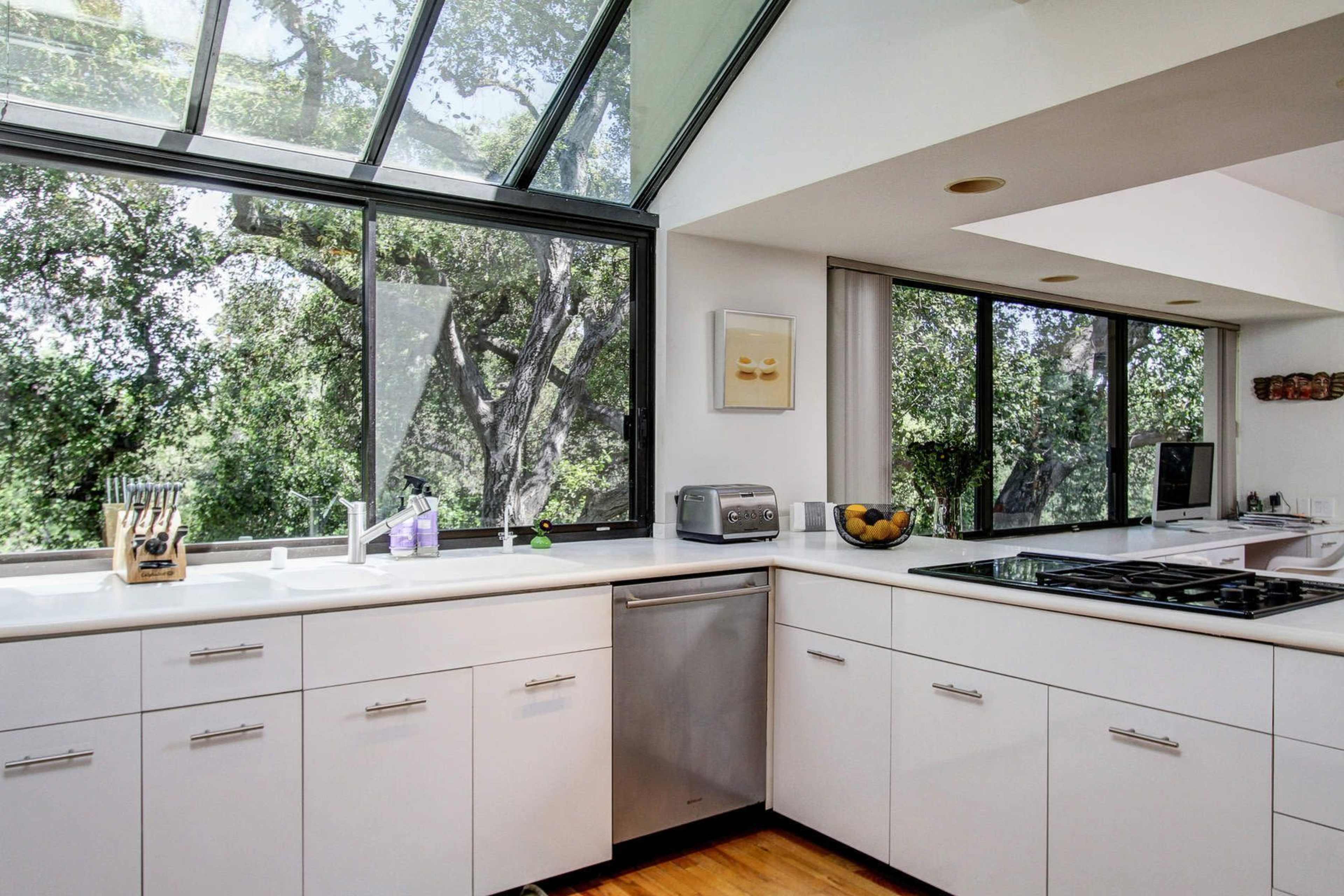 A modern kitchen features white cabinetry, a stainless steel dishwasher, and large windows overlooking a green landscape.