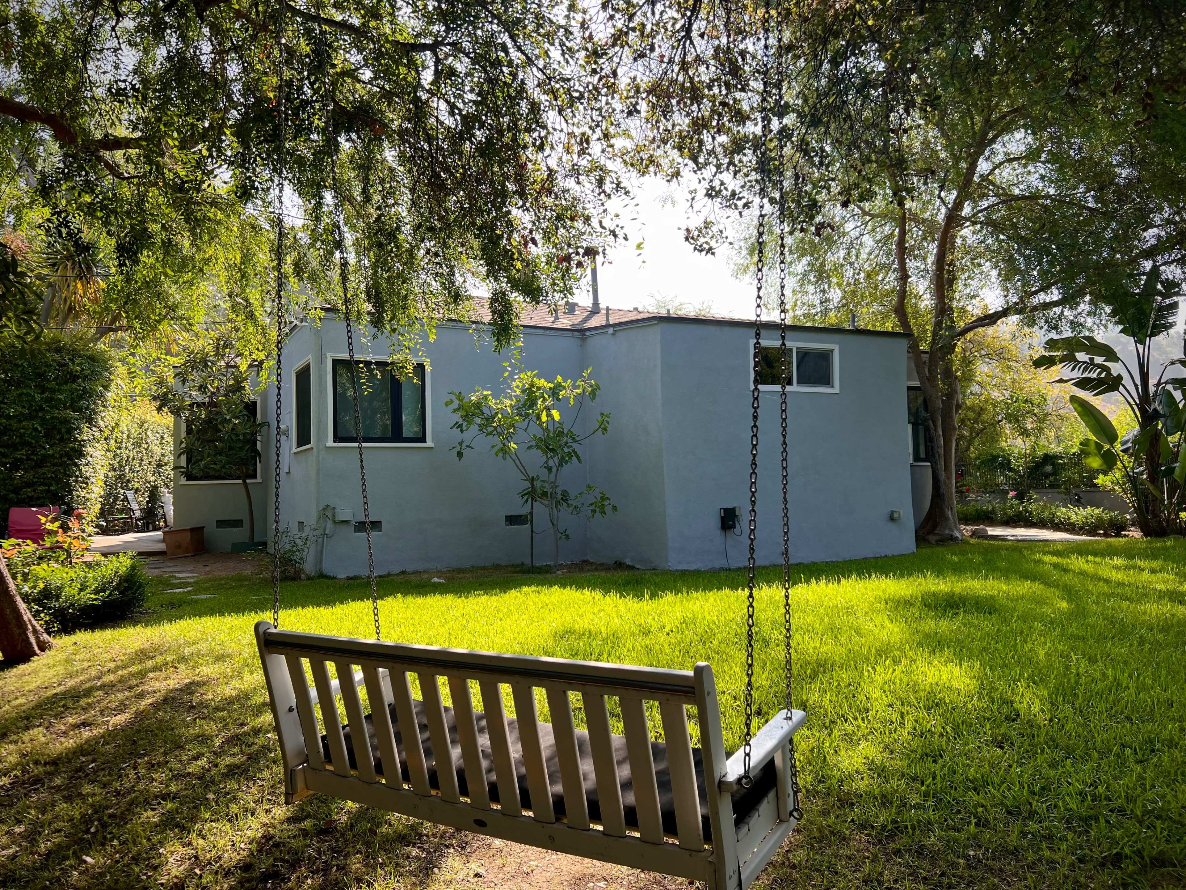A wooden swing hangs in the foreground of a backyard, facing a pale gray house surrounded by lush greenery.