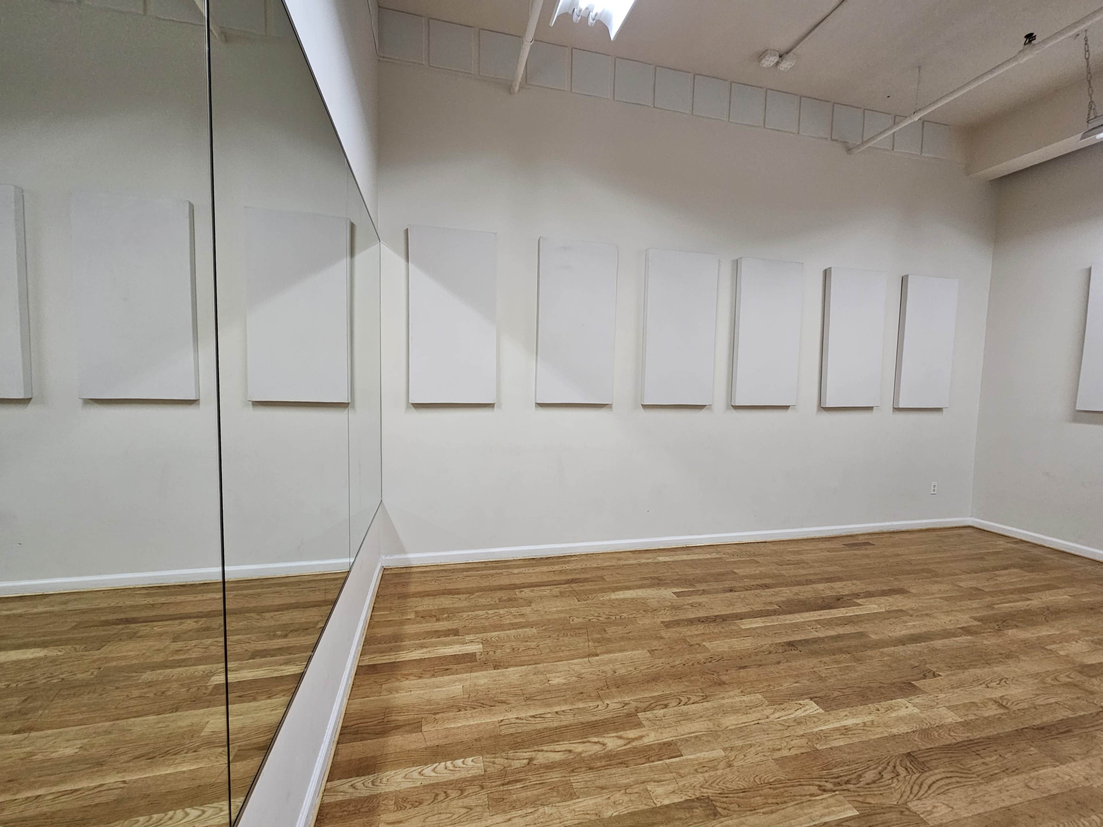 An empty room with a wooden floor, white walls, and eight blank white canvases mounted on one wall, along with a large mirror reflecting the opposite wall.