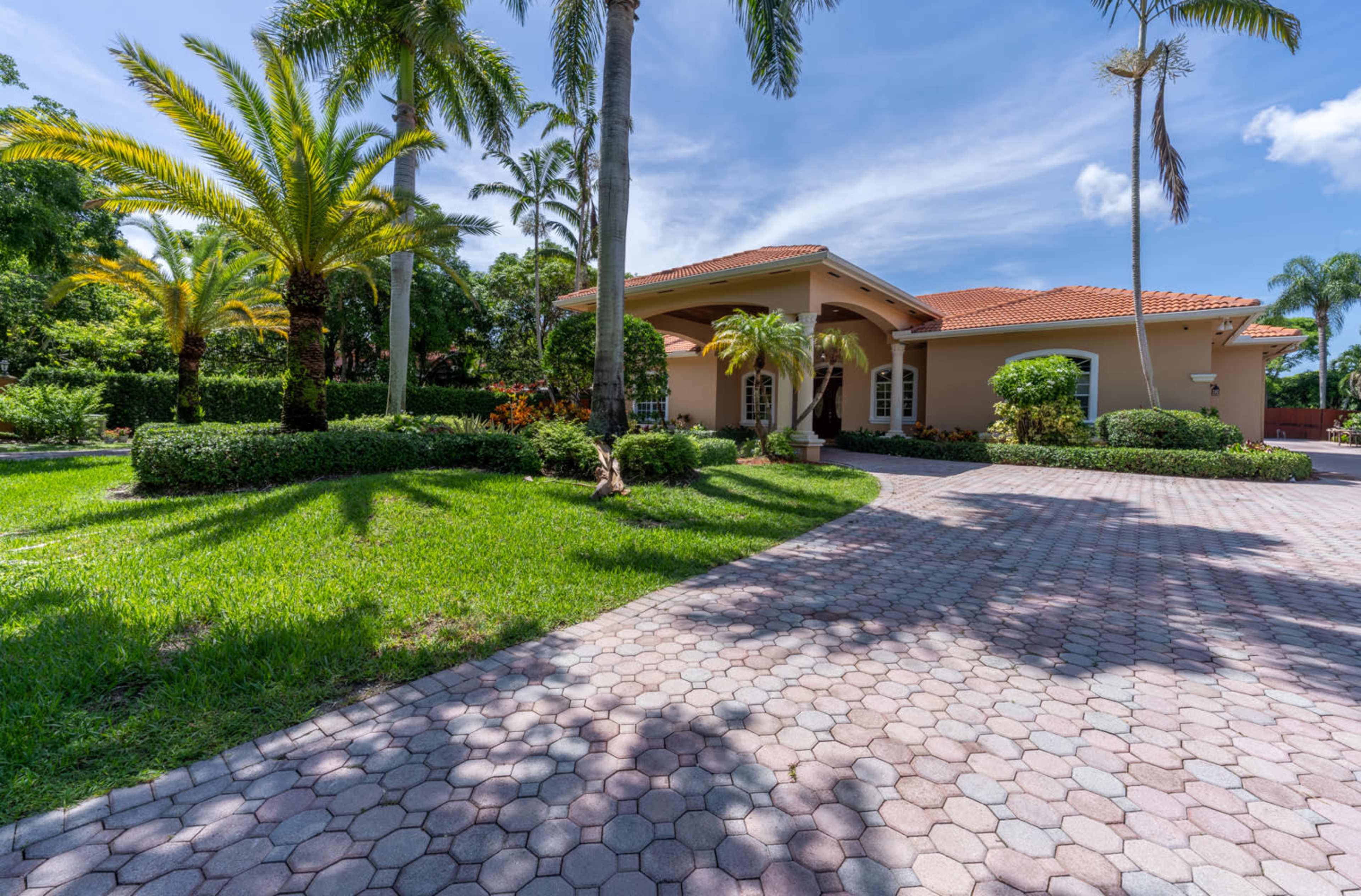 A single-story house with a red-tiled roof is surrounded by palm trees and well-maintained landscaping, with a brick driveway leading to the entrance.