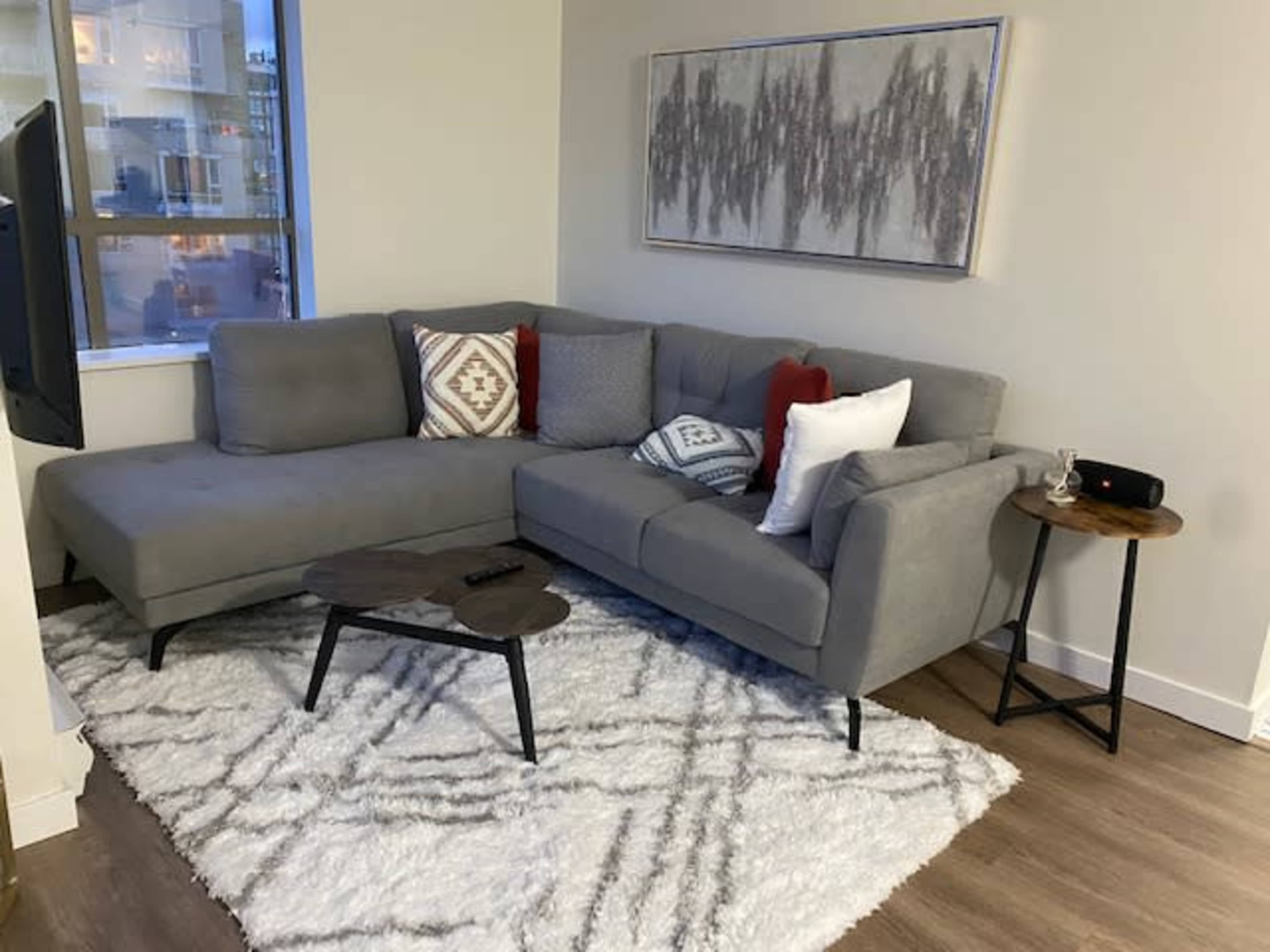 A gray sectional sofa with red and white accent pillows sits on a patterned rug beside a small wooden side table in a well-lit room.