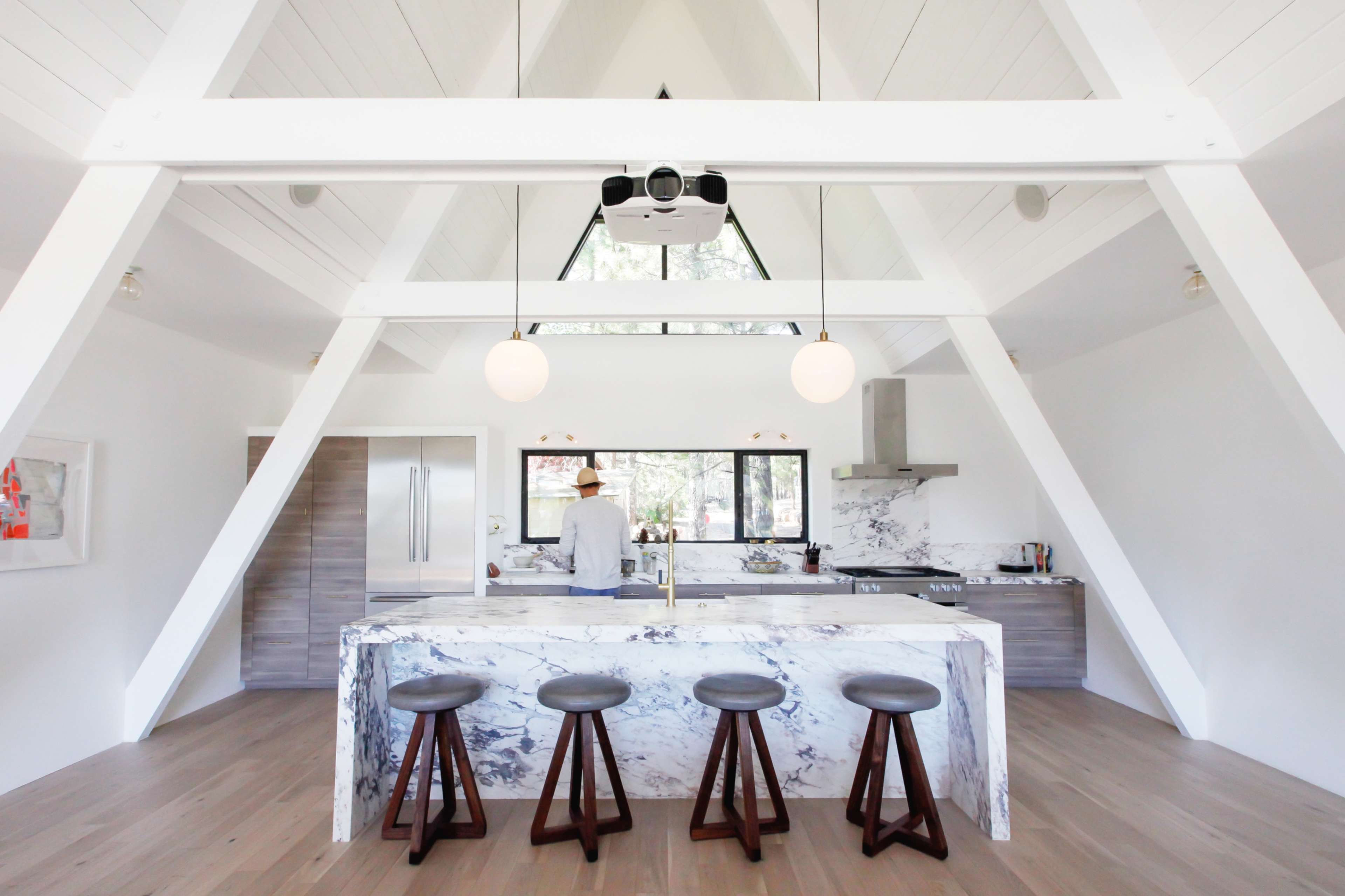 A modern kitchen with a marble island and four wooden stools, featuring high ceilings and large windows that provide natural light.