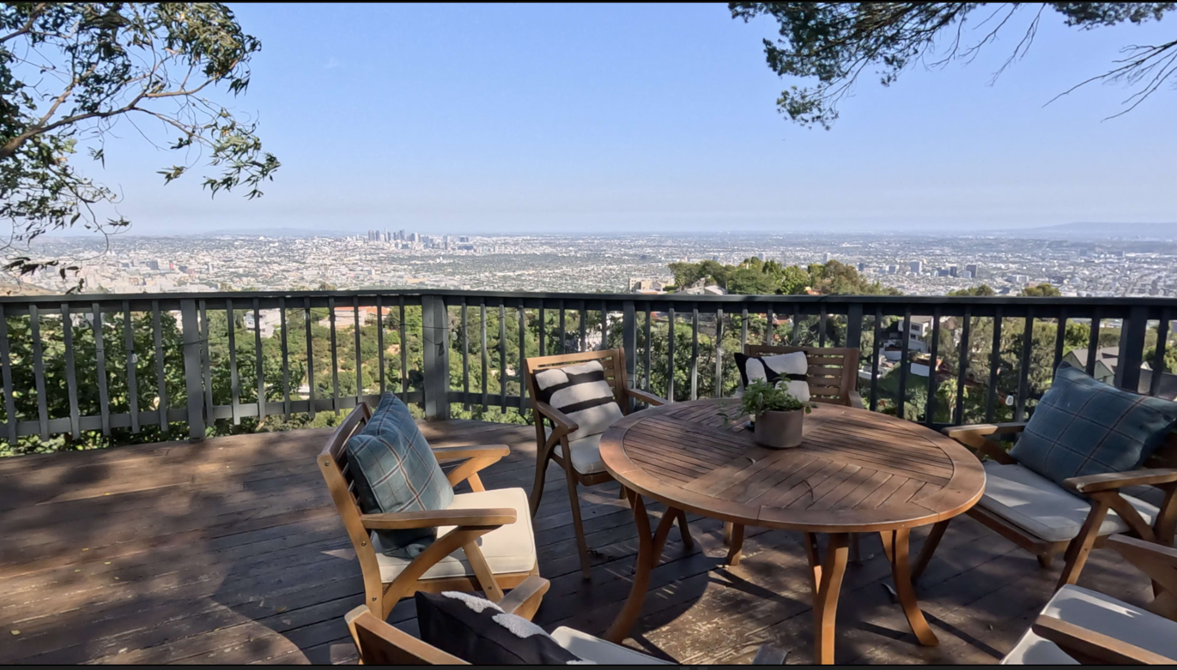 A circular wooden table surrounded by several chairs is set on a deck, offering a panoramic view of a city skyline in the distance.