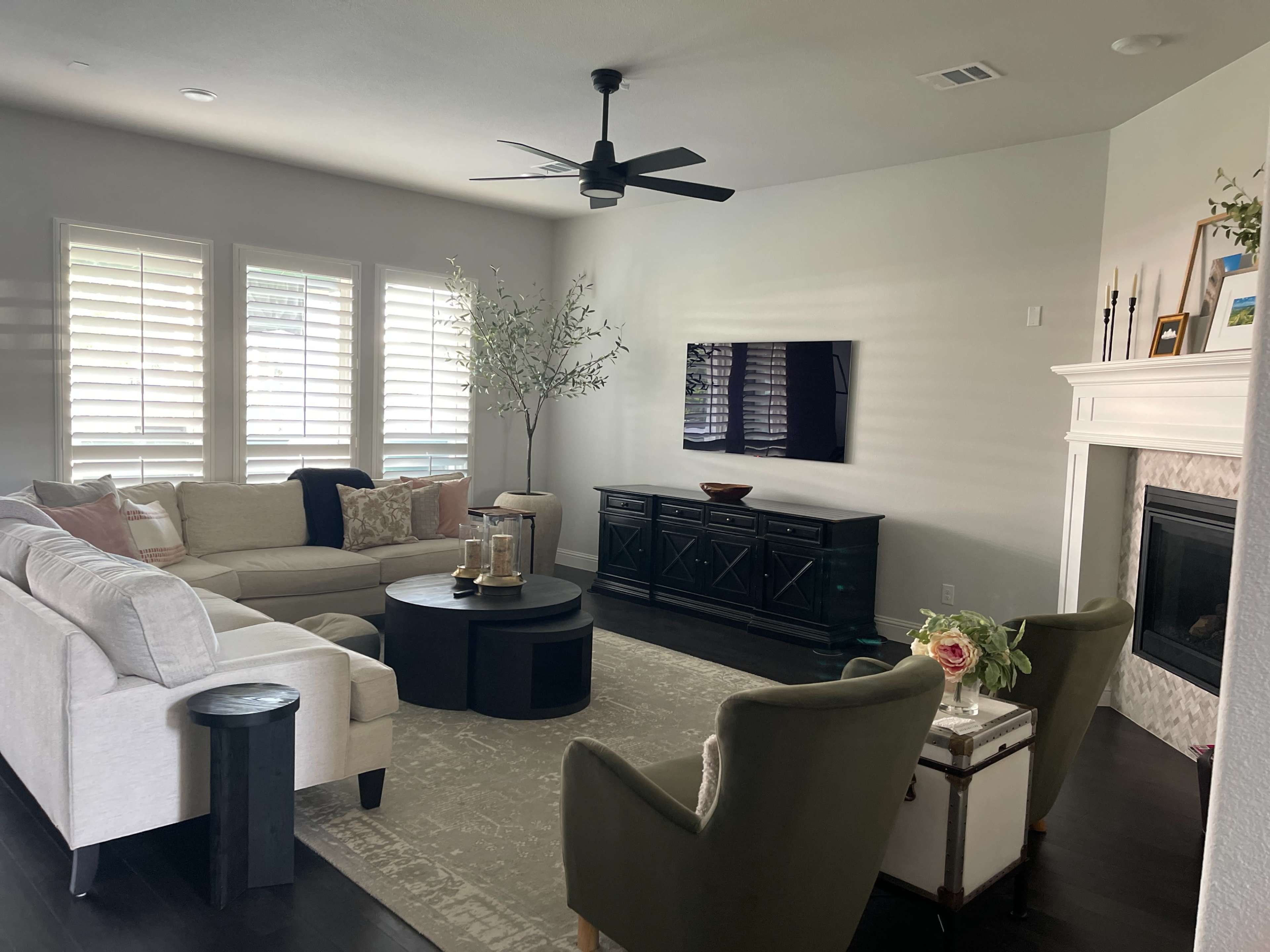 The living room features a light-colored sectional sofa, a round coffee table, a television mounted on the wall, and a mix of decorative elements, such as a plant and flowers.