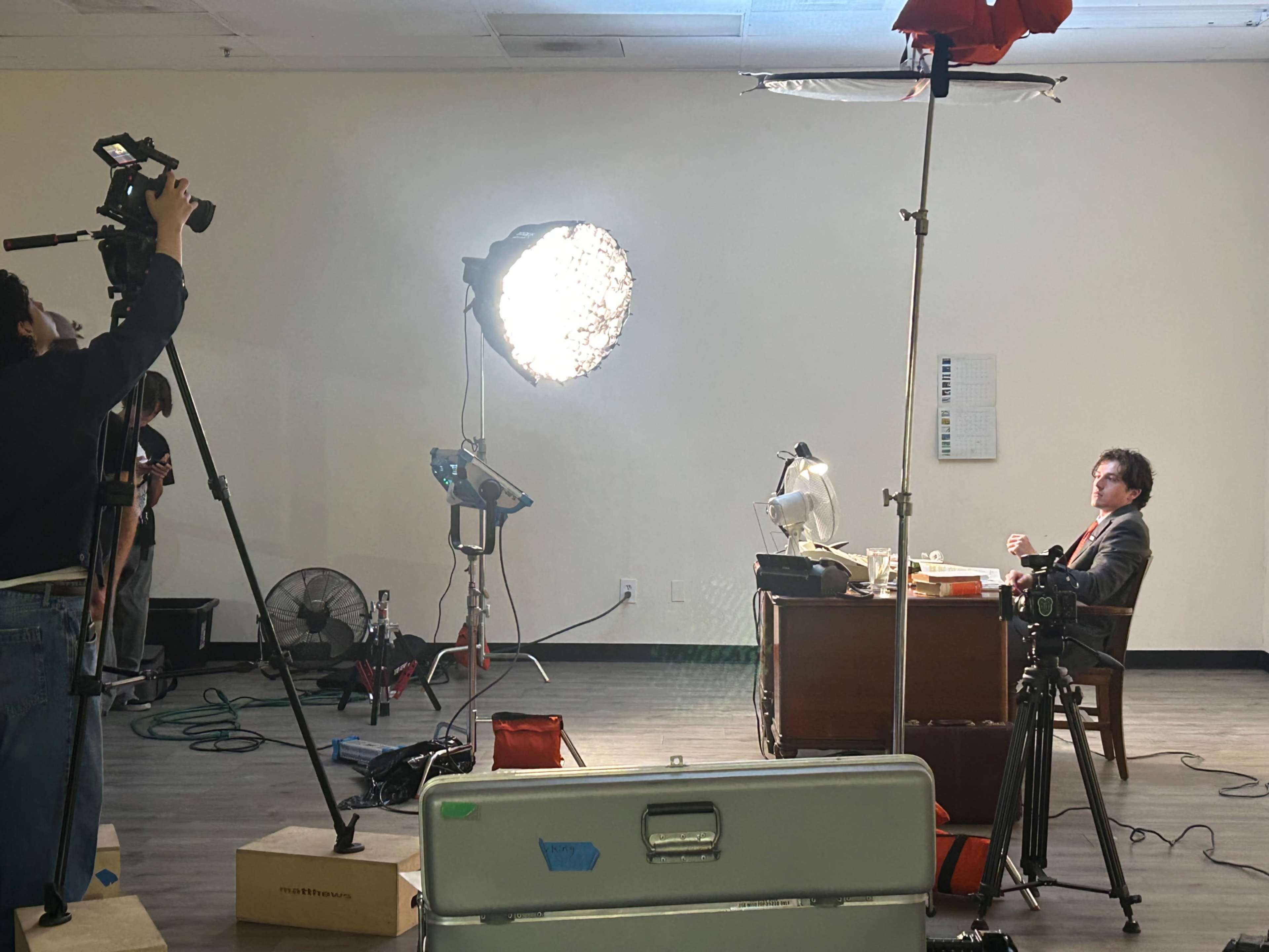 A film crew is set up in a well-lit room, with one person operating a camera, while another sits at a desk with various items in front of them.