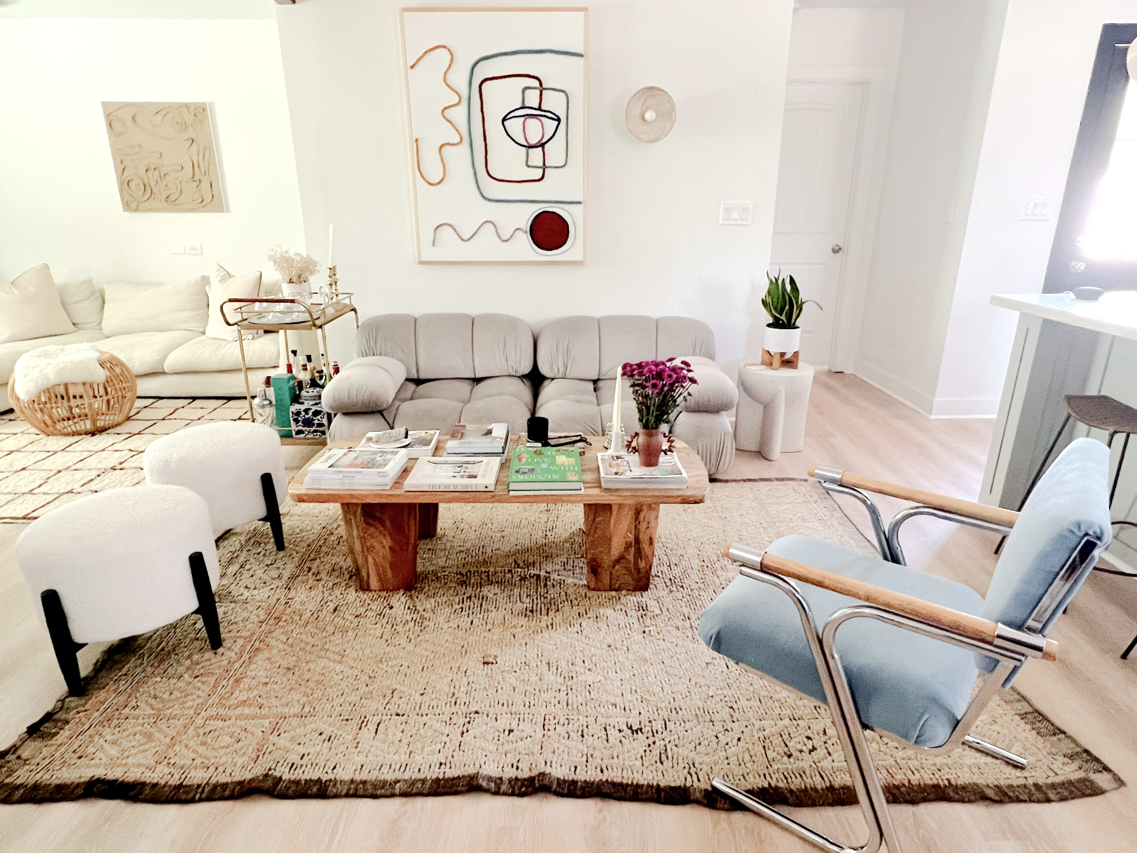 The image shows a modern living room featuring a light-colored sofa, a wooden coffee table with magazines, two minimalist armchairs, and decorative art on the walls.