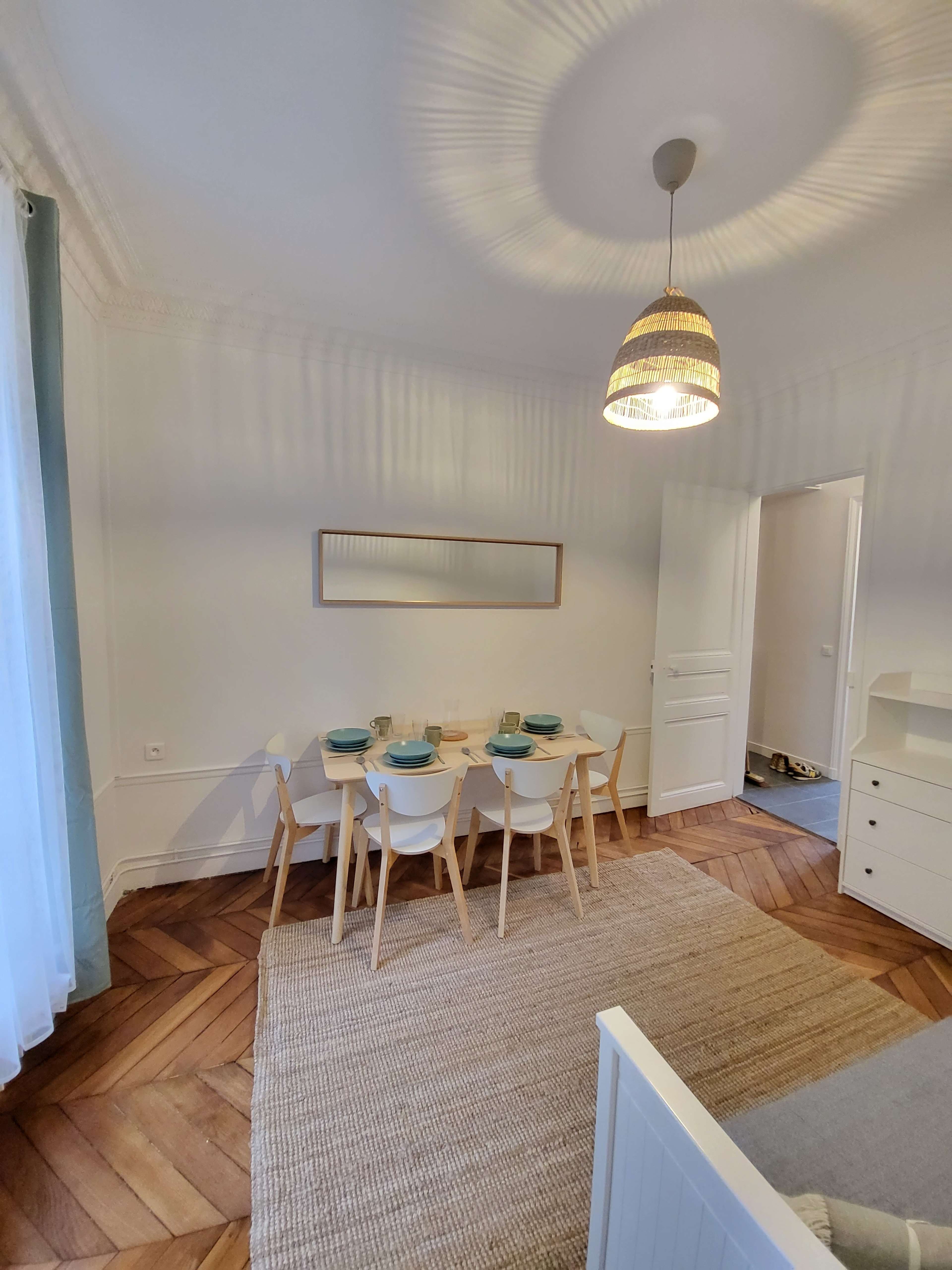 A dining area with a light-colored table and four chairs is set against a wall with a framed picture, illuminated by a pendant light.