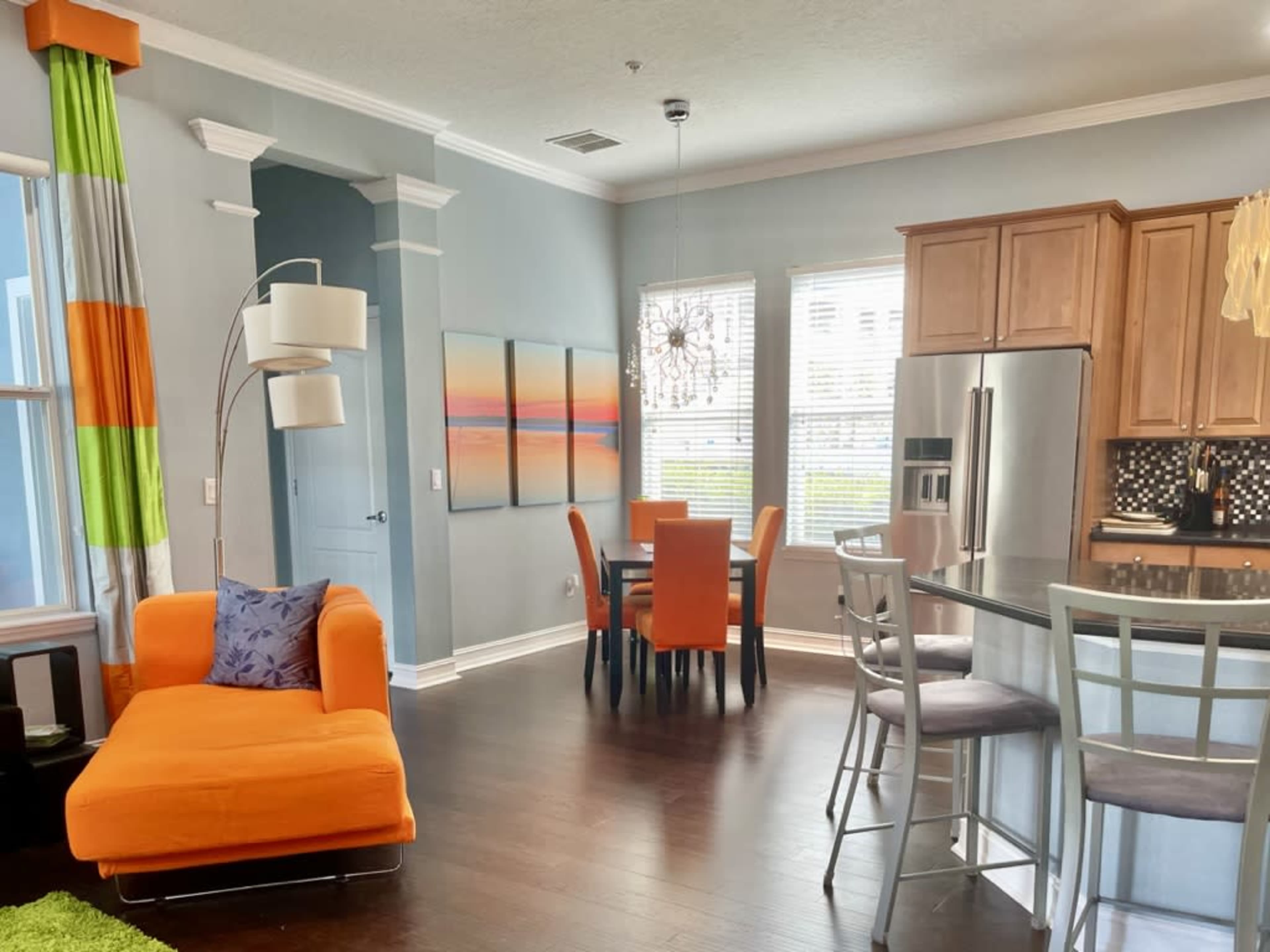 A modern living space with an orange sofa, a dining area with orange and gray chairs, and a kitchen with wooden cabinets and stainless steel appliances.