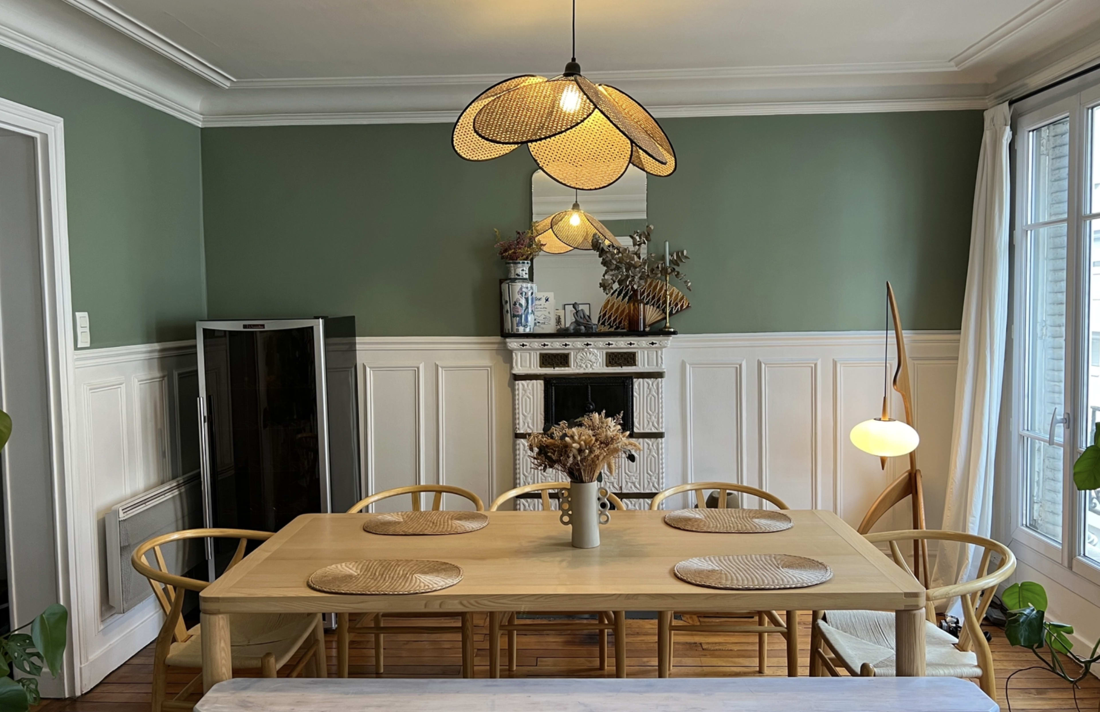 The image shows a dining area with a wooden table surrounded by chairs, a decorative mirror, and light fixtures against a green wall.