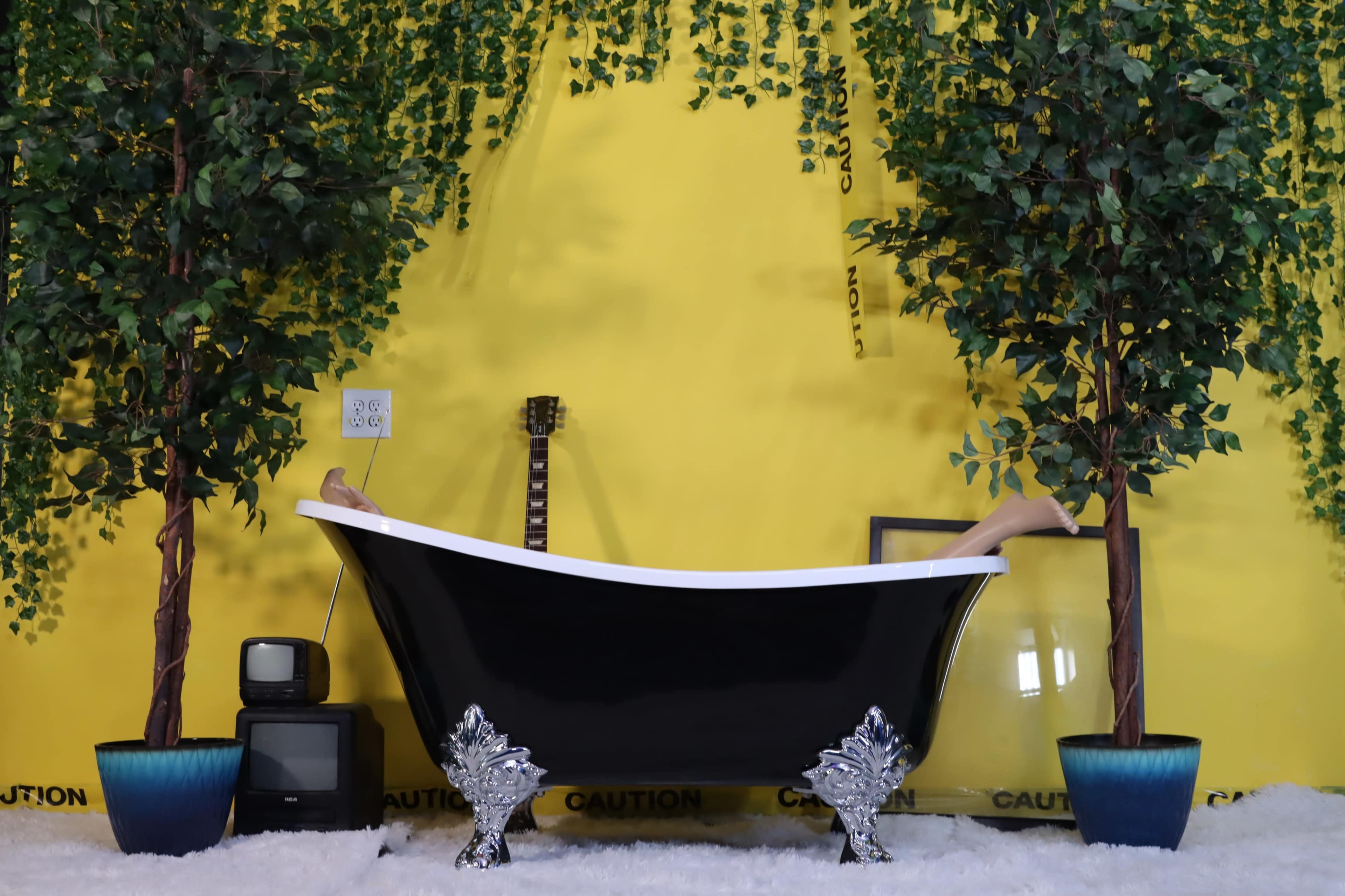 A black clawfoot bathtub is centered against a bright yellow wall, flanked by two artificial trees and a vintage television, with a guitar leaning nearby.