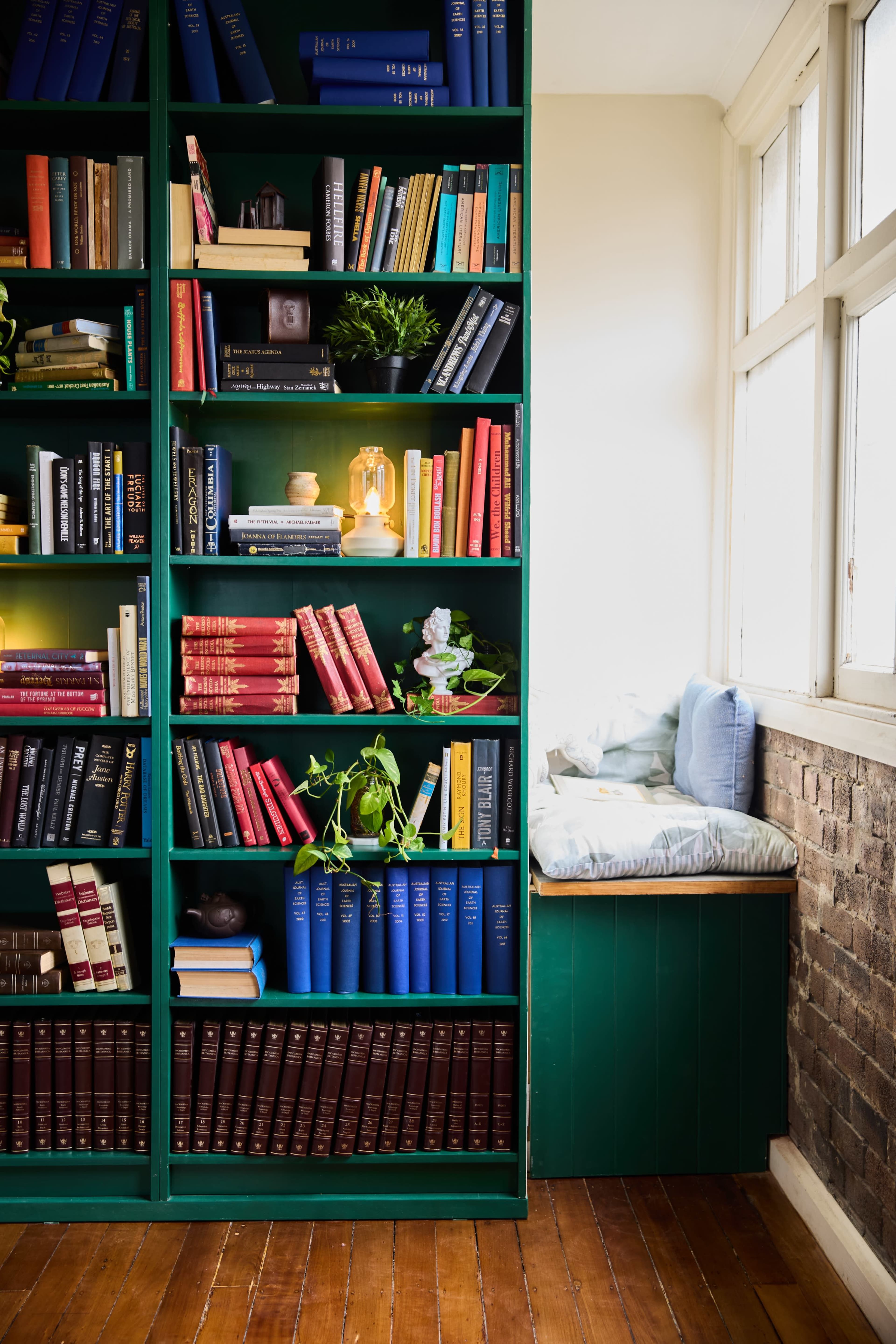 The image features a tall green bookshelf filled with a variety of books, accompanied by decorative plants, while a cozy window seat is visible beside it.