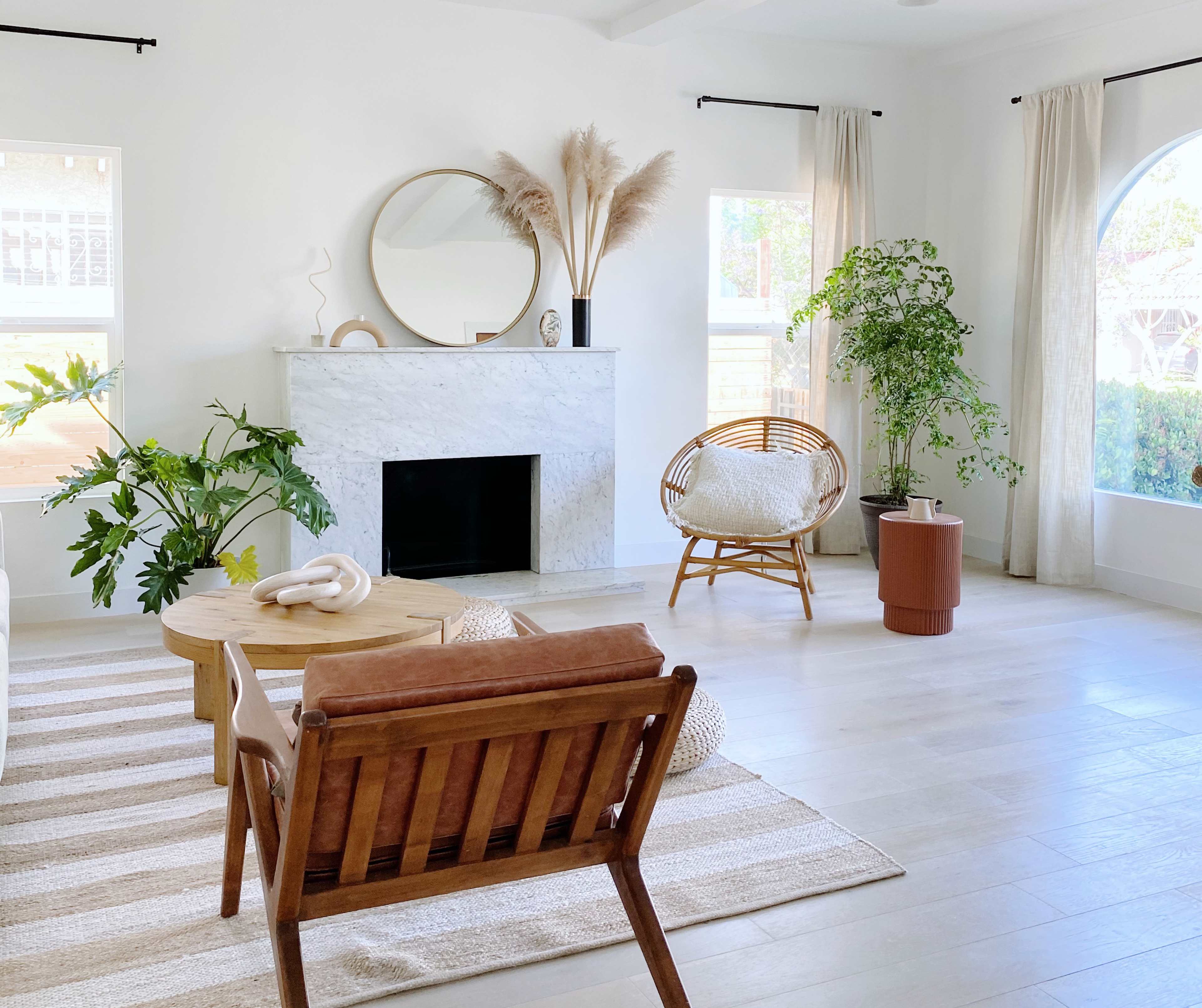 A modern living room with a white stone fireplace, a round mirror, a rattan chair, and various plants.