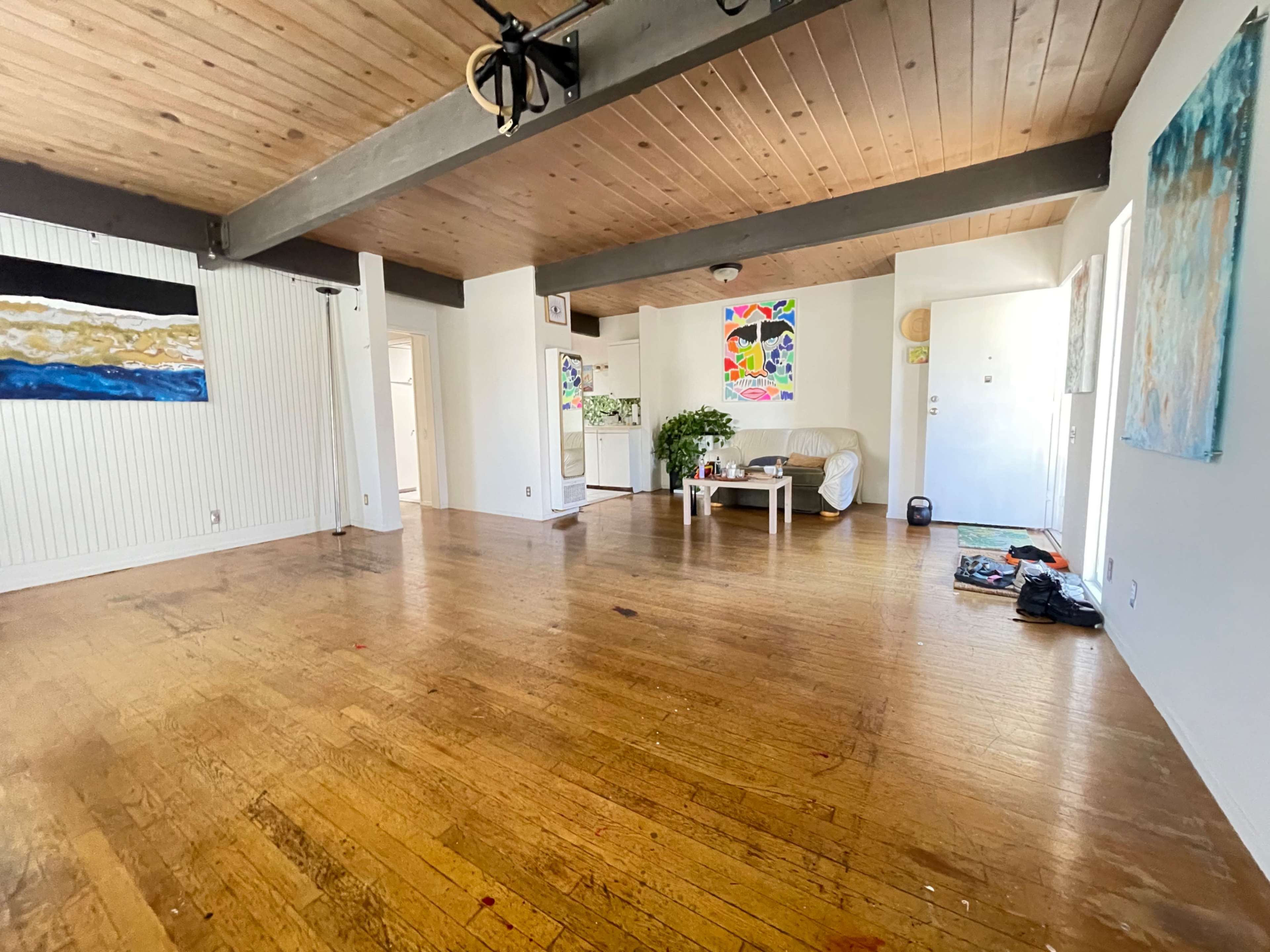 A spacious, empty living room with wooden floors, large windows, a beige couch, and colorful artwork on the walls.