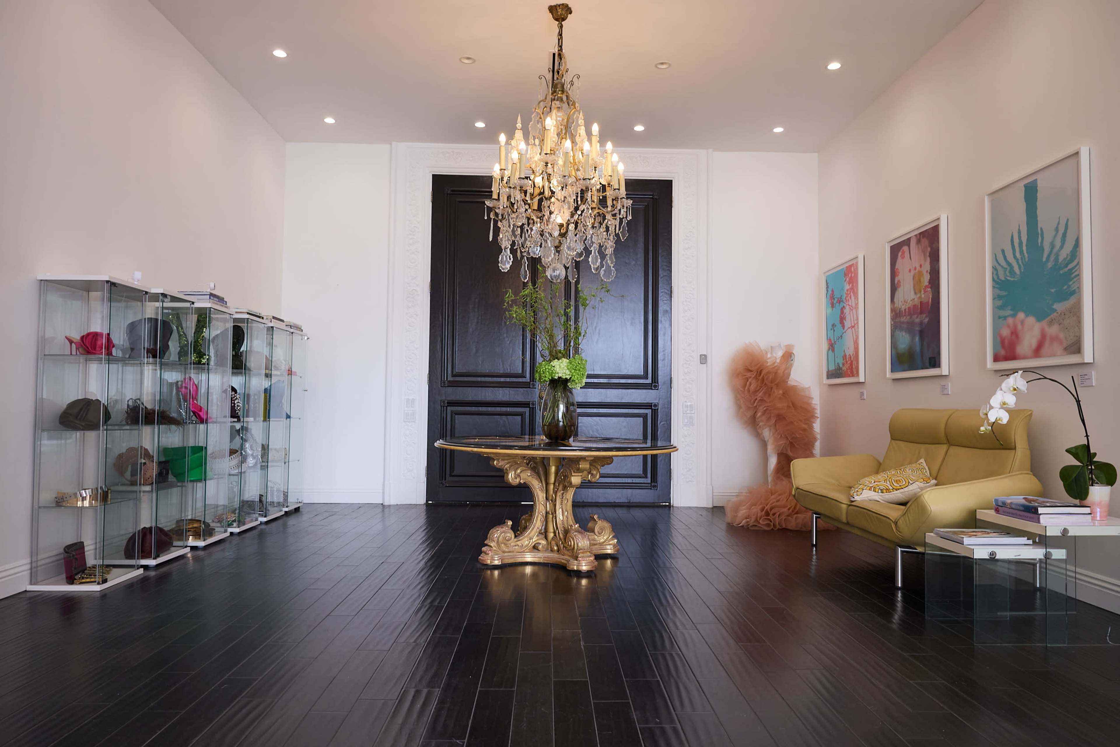The image shows a well-lit foyer featuring a large black double door, a golden circular table with a vase of flowers, a chandelier overhead, a plush light-colored sofa, and a display case filled with colorful accessories.