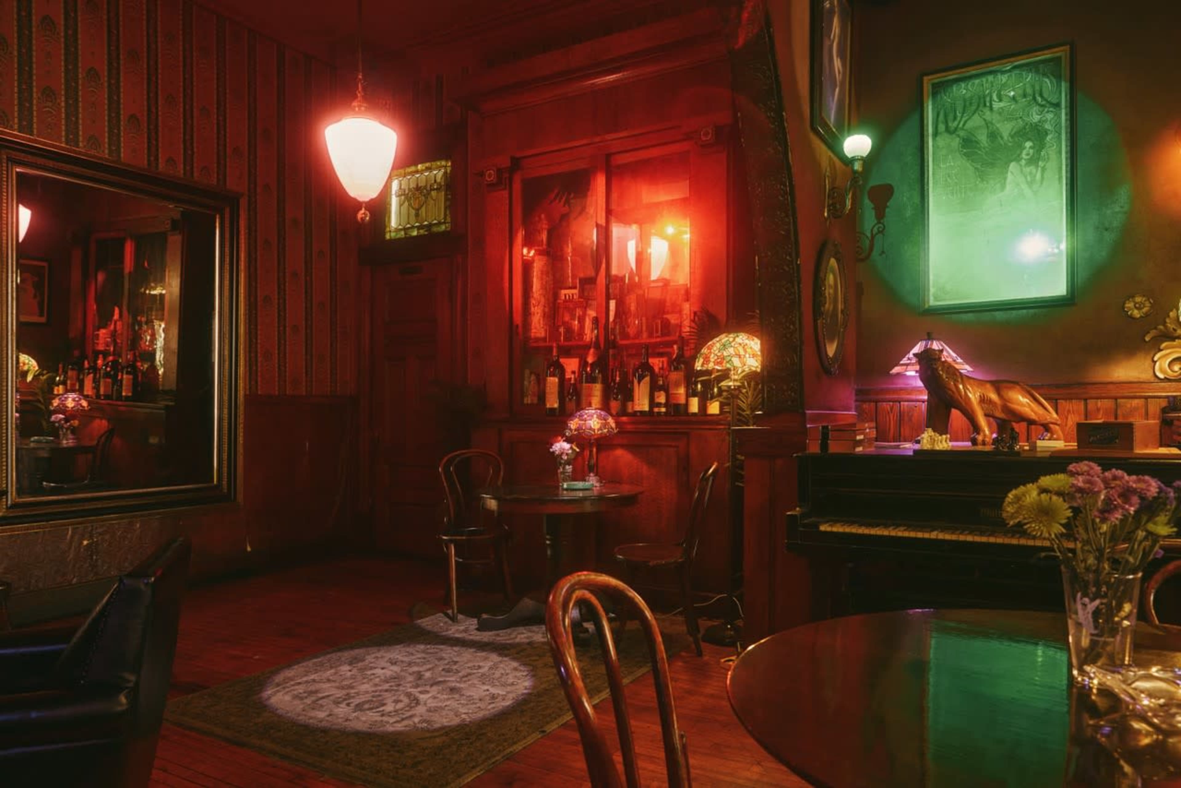 The image depicts a dimly lit vintage bar featuring a round table with flowers, a piano, an ornate mirror, and various bottles displayed on shelves against a red-lit wall.