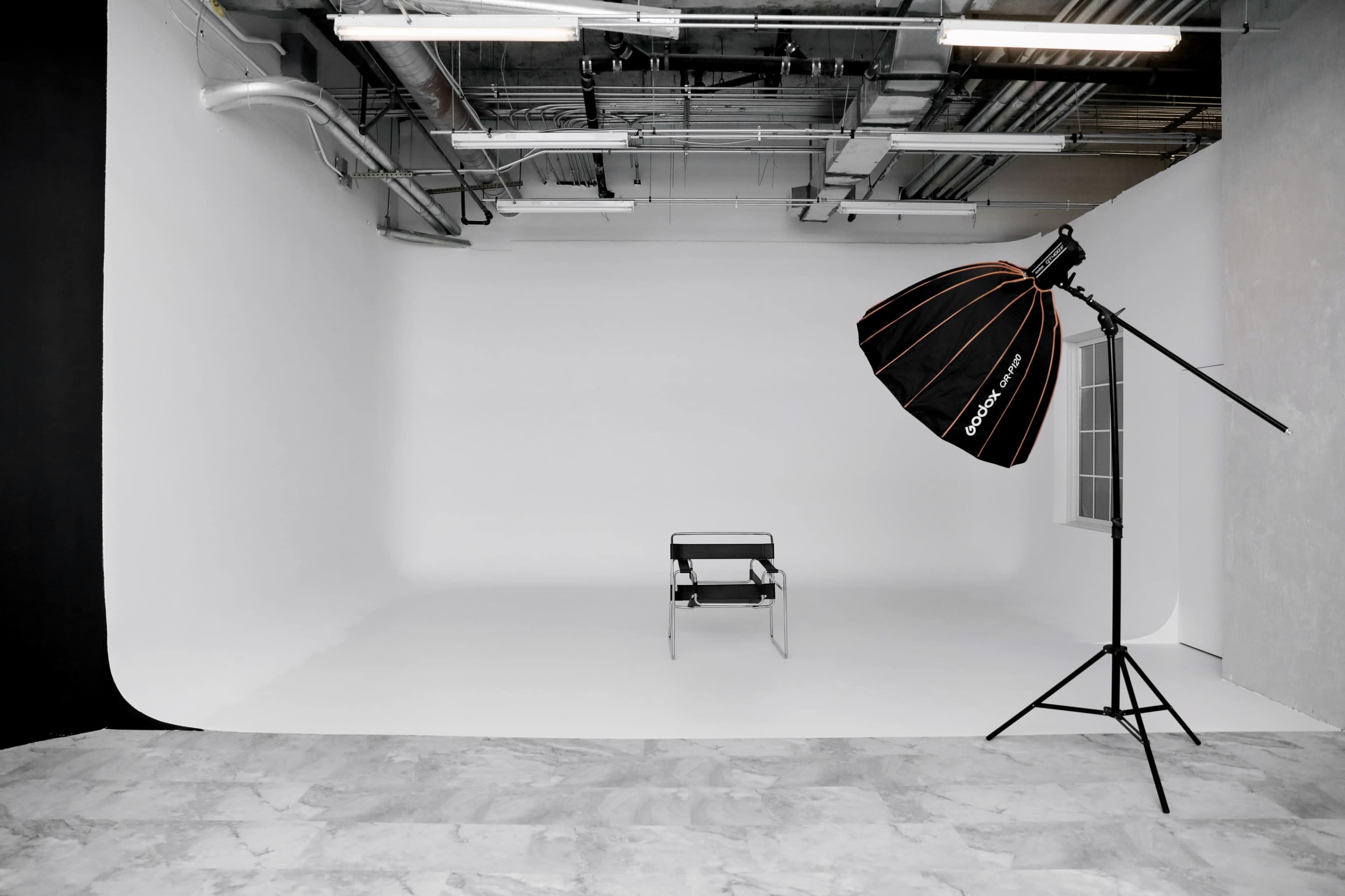 A minimalist photography studio with a single black chair and a large softbox lighting setup against a white backdrop.