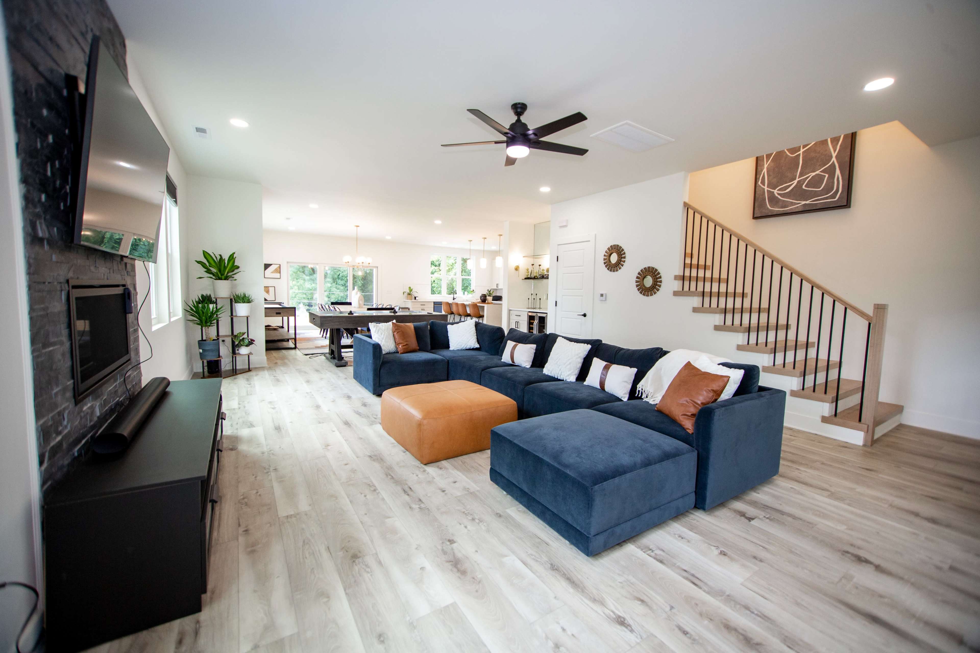 A modern living room features a large blue sectional sofa, a flat-screen TV mounted on the wall, and a staircase leading to an upper level.